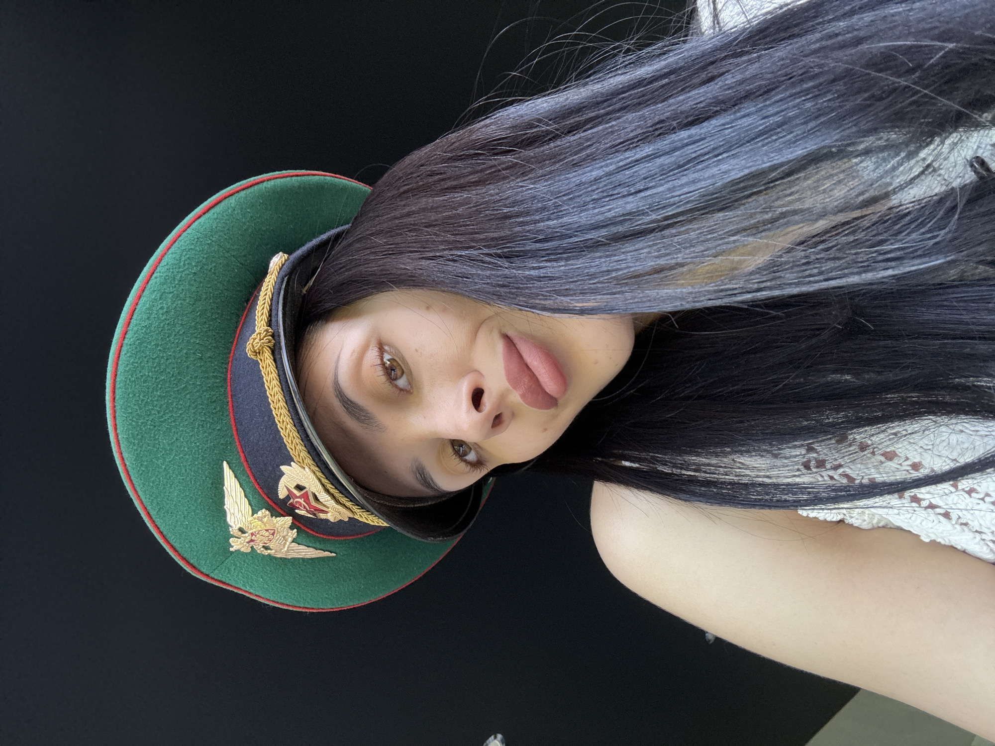 A young woman with long, dark hair is wearing a green military-style cap with gold embellishments. She is sticking her tongue out slightly and looking directly at the camera. The background is black. Part of a white, lacy garment is visible on her shoulder. There is no evidence to suggest the location is Votkinsk, Russia.