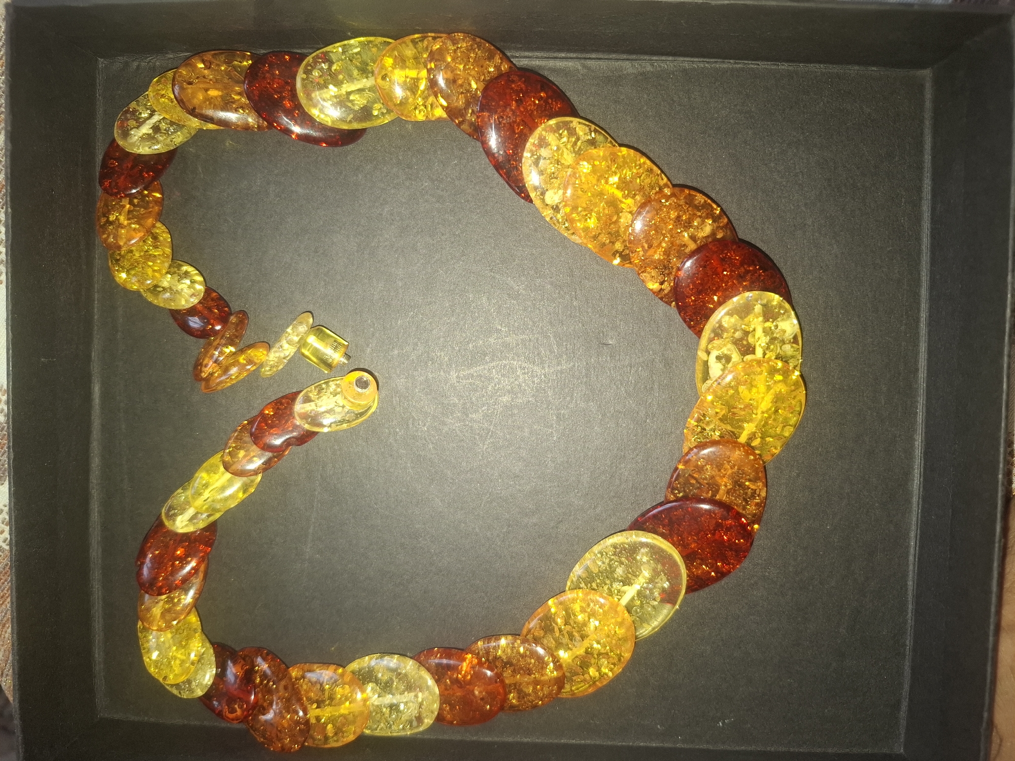 A heart-shaped necklace made of oval amber beads in various shades of yellow and red is displayed in a dark rectangular box. The necklace is coiled to form the heart shape. The background is a dark, textured surface, possibly fabric, with a lighter border at the top. The scene is indoors.