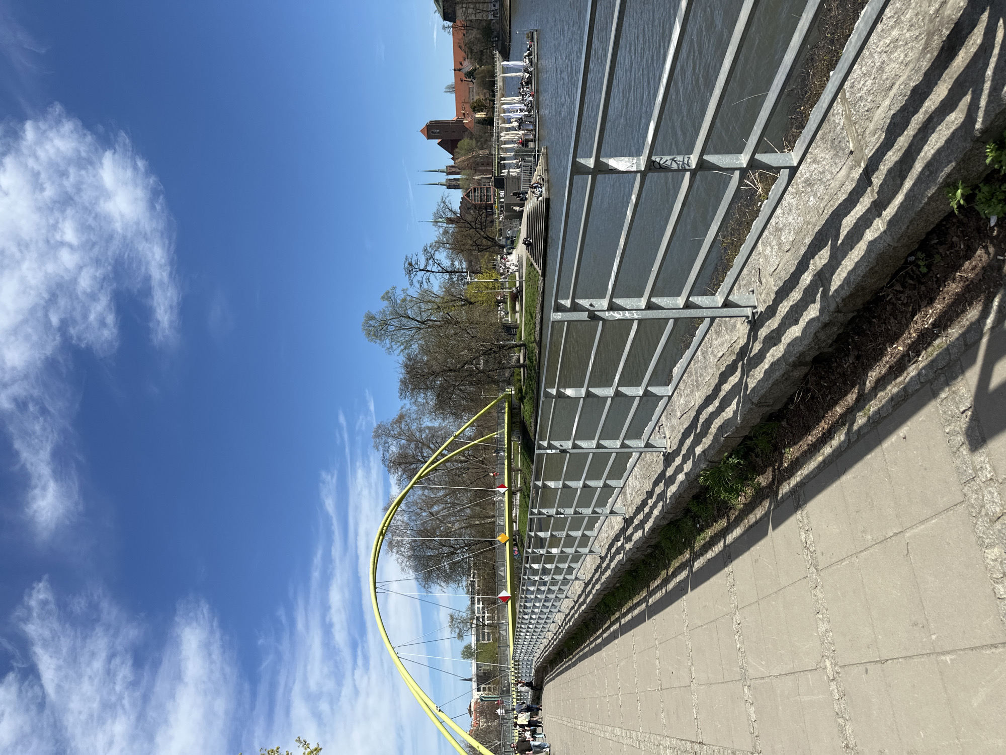 The image depicts a sunny day alongside the Oder River in Wrocław, Poland. A pedestrian walkway runs along the river's edge, with a metal railing separating it from the water. The walkway appears to be constructed of concrete slabs with stone borders.
A bright yellow suspension bridge, known as Most Tumski, arches over the river. Buildings, including the spires of churches, are visible in the background across the river.
People are present on the walkway, suggesting a public space for recreation. Shadows cast by the railing indicate the direction of the sunlight. Vegetation is sparse along the edge of the walkway, growing between the railing and the river bank. The sky is blue with scattered white clouds.