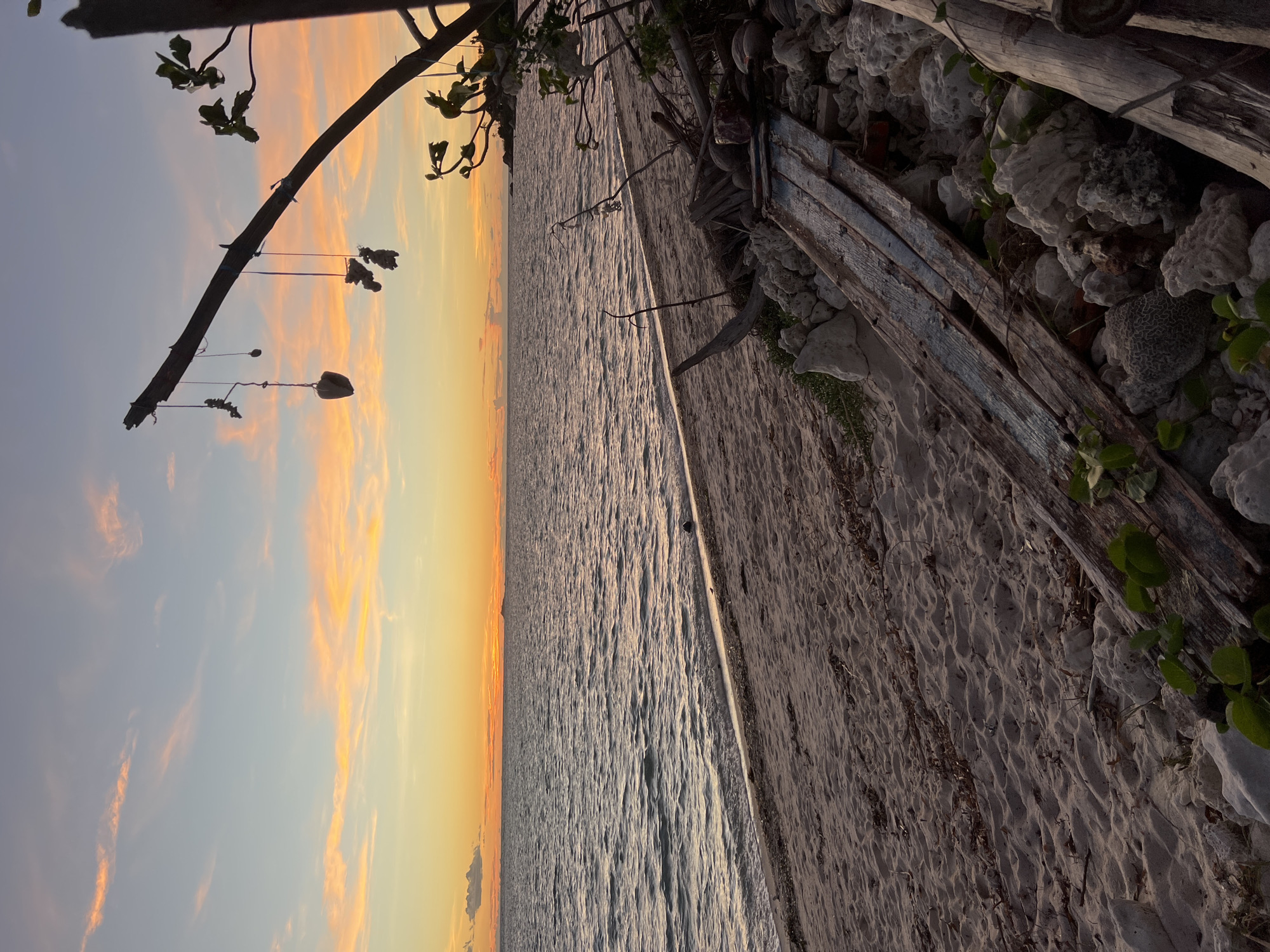 This is a vertical, eye-level shot of a beach at sunset on Koh Phangan, Thailand. The sky is a gradient of pale blue at the top, transitioning to soft orange and yellow hues near the horizon, with wispy clouds tinged pink. The ocean is calm with gentle ripples reflecting the sunset colors. The beach is sandy with scattered pieces of driftwood, rocks, and some vegetation. In the foreground, to the right, is a pile of rough-hewn wood and coral rocks, with a couple of weathered wooden planks leaning against them. A vertical wooden pole, silhouetted against the sky, extends from the right side of the frame towards the center, with three objects suspended from it by strings. The one closest to the pole appears to be a small, light-colored rock, followed by two darker, irregularly shaped objects that could be shells or small stones. The overall atmosphere is serene and natural.