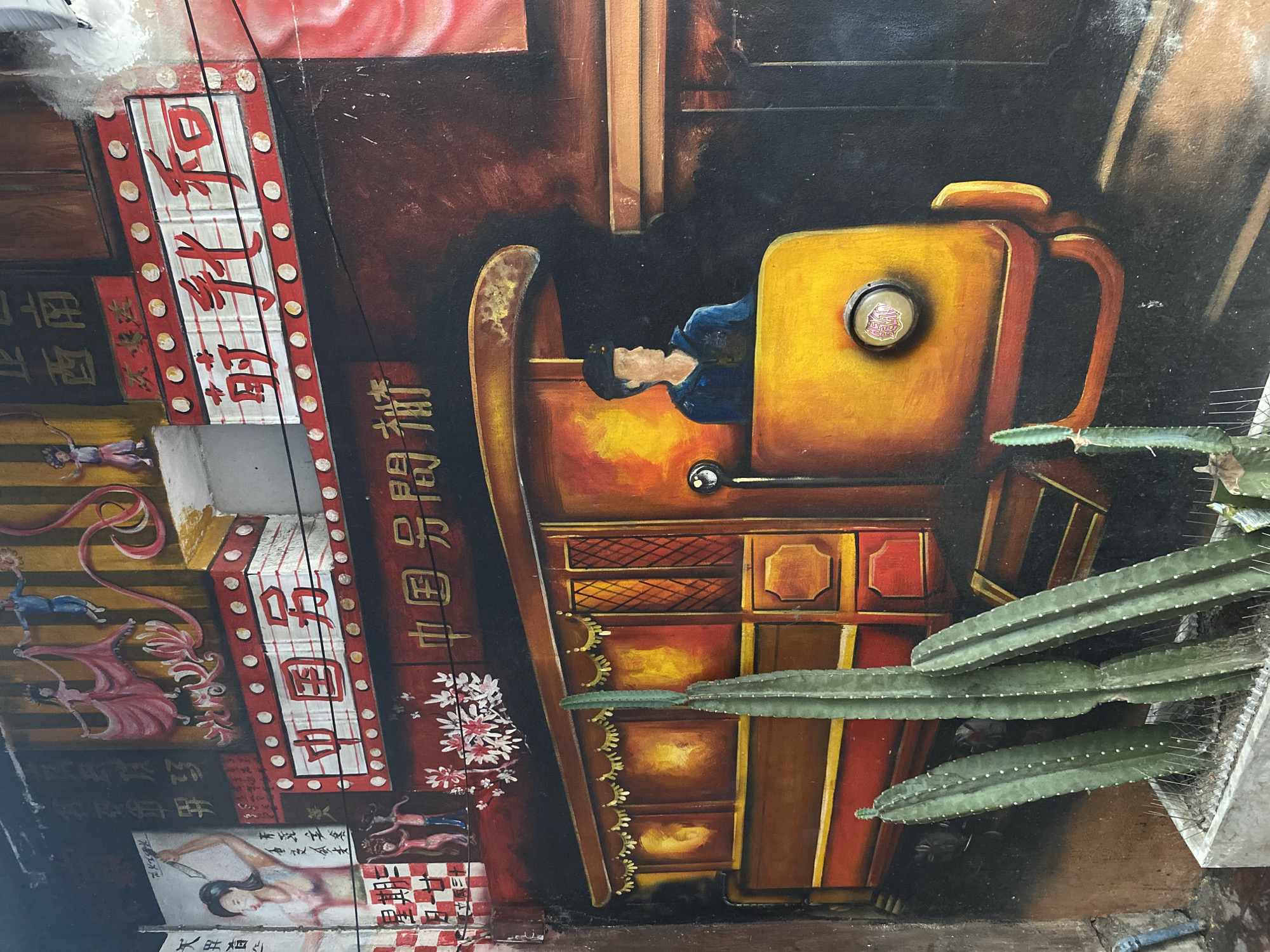 The image shows a mural in Bangkok, Thailand. The mural features a retro-style illustration of a man operating a machine. The mural incorporates Chinese text and decorative elements. A cactus plant is positioned in front of the mural, partially obscuring the lower right corner. The artwork appears to be part of a larger installation or street art project.