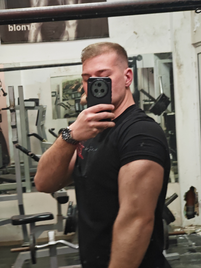 A male individual is depicted taking a mirror selfie within a gym environment in Shumen, Bulgaria. The subject, with short, faded light-colored hair, is wearing a black short-sleeved t-shirt. He holds a black smartphone with a distinctive multi-lens camera system in his right hand, obscuring a portion of his face from the eyes upward. On his left wrist, he wears a dark-colored wristwatch with a metal band and a red accessory, possibly a string or bracelet. His left arm is flexed, displaying muscle definition.
The background, visible as a reflection in the mirror, shows various pieces of gym equipment, including metallic frames, a weight bench with a black padded seat, and weight racks. Above the subject's head in the reflection, a poster partially displays the name "Arnold." The walls of the facility are light-colored, showing visible signs of wear, including peeling paint and an irregular area of damage on the right side of the frame. Another framed image depicting gym equipment is visible further in the reflection. The scene is illuminated by interior lighting.