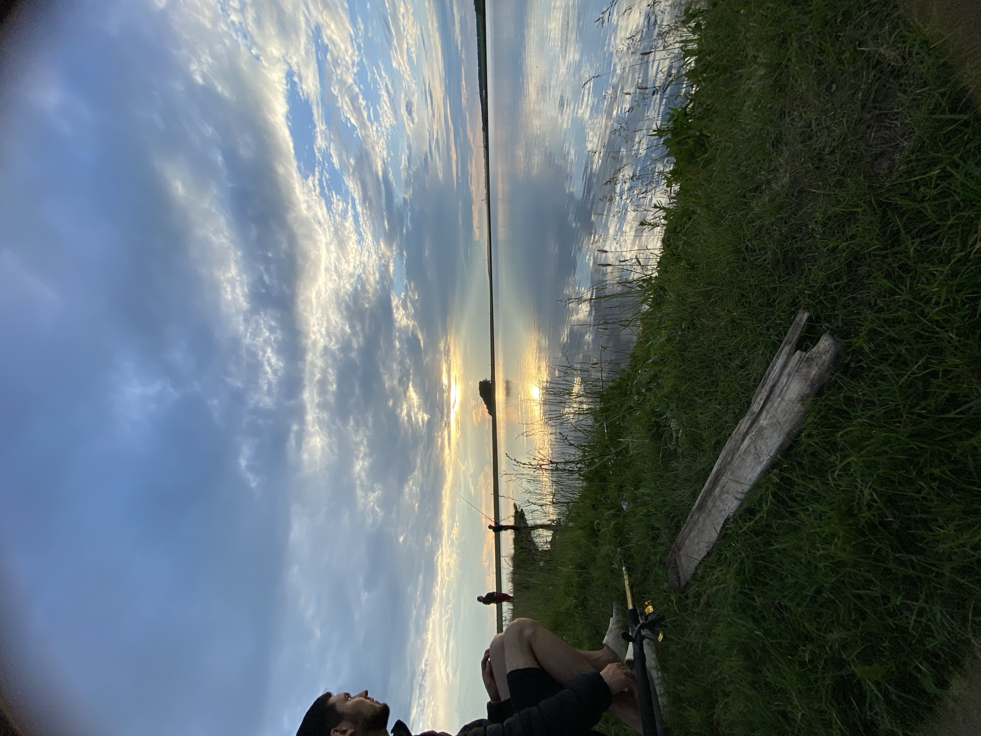 In Сыйрышбашево, Russia, a man crouches on the grassy bank of a calm body of water, looking towards a sunset. The sky is filled with dramatic clouds, reflecting on the water's surface. Two other figures are visible in the distance, also by the water's edge, appearing to be fishing. Fishing rods are present in the foreground near the crouching man. The scene suggests a peaceful evening by the water.