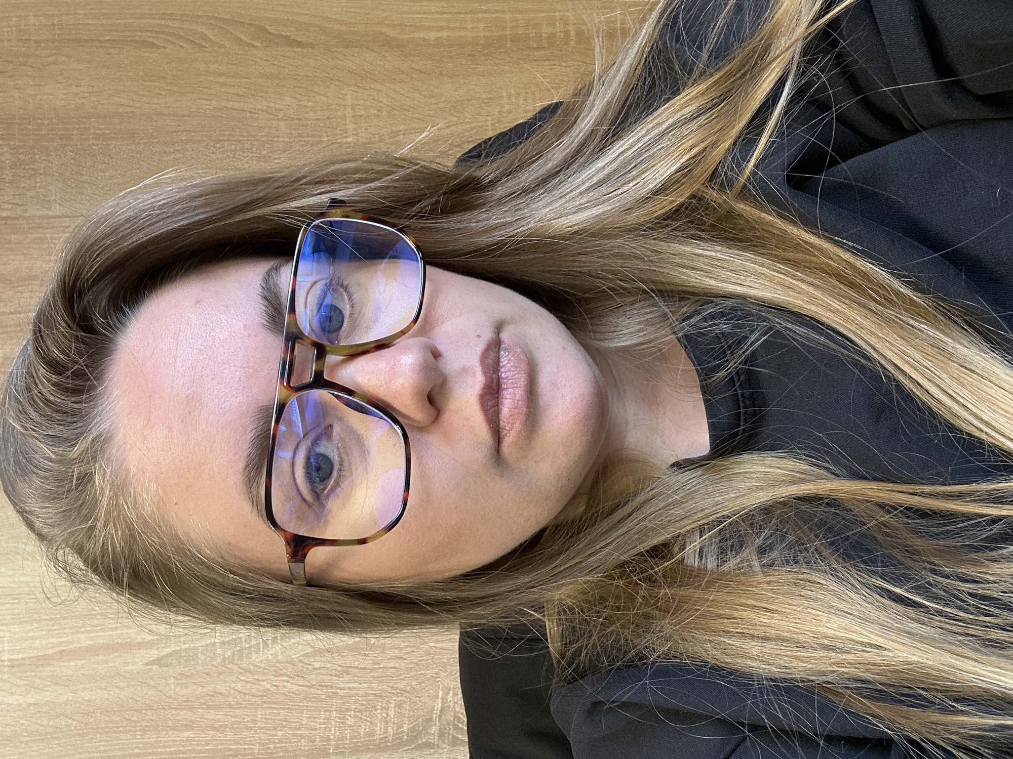 A woman is wearing eyeglasses and a black top. She has long, blonde hair and is lying on a light brown wooden surface. The location is not discernible from the image and cannot be determined.