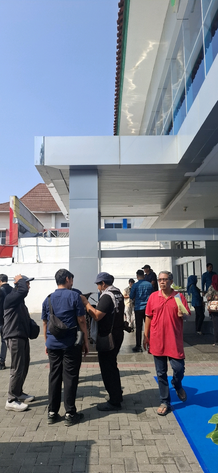 The image shows a group of people standing outside a modern building with a large glass facade and a covered walkway. The ground is paved with interlocking brick pavers. In the foreground, three men are standing. The man on the left is wearing a black hooded jacket and dark pants. The man in the middle is wearing a blue t-shirt and dark pants with a black crossbody bag. The man on the right is wearing a bright red polo shirt and blue jeans, holding a yellow piece of paper. There are other individuals visible in the background, some walking and some standing. A blue mat is present on the right side of the image. The overall scene suggests an outdoor gathering or event. The building's architecture indicates a contemporary design. The clear blue sky suggests it is daytime. The image does not contain any specific elements that would pinpoint the exact location within the city of Surakarta, Indonesia, other than it is an outdoor urban setting.