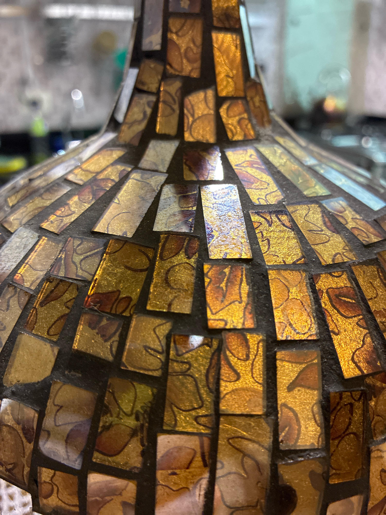 The image shows a close-up view of a mosaic pattern on a vase or decorative object. The mosaic is made of small, rectangular tiles in shades of gold, amber, and brown. Some of the tiles have a textured, slightly iridescent finish, while others appear to have a subtle floral or abstract pattern etched into them. The tiles are set against a dark grout. The background is blurred, suggesting it is out of focus, but hints at an indoor environment with some objects and possibly lighting visible. There are no people or animals present in the image, nor any identifiable landmarks or features that would indicate a specific location in Curitiba, Brazil. The focus is entirely on the detailed texture and colors of the mosaic.