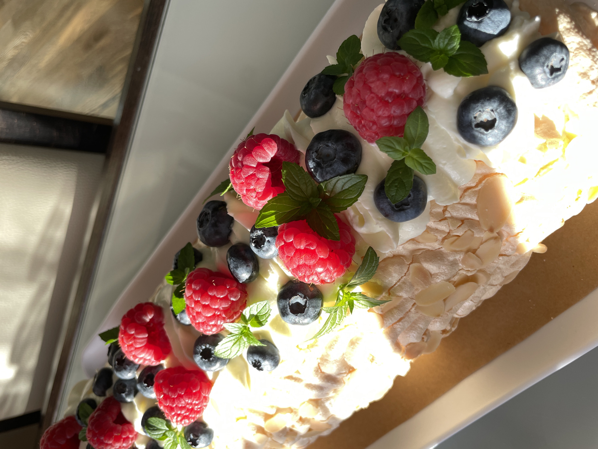 The image shows a close-up view of a meringue roulade dessert, adorned with whipped cream, fresh raspberries, blueberries, and mint leaves. The dessert is presented on a white rectangular platter, which is placed on a light brown paper liner. The platter is positioned on a white surface, likely a table or countertop. Natural light streams in from the right side of the frame, casting shadows and highlighting the textures of the dessert and its toppings. The background is out of focus, but appears to be an indoor setting with hints of neutral-colored walls and possibly a window frame or furniture. There are no people or animals visible in the image, and no action is taking place. The scene is static and focused solely on the dessert. The location in the city of Saint Petersburg, Russia cannot be determined from the visual information provided in the image.