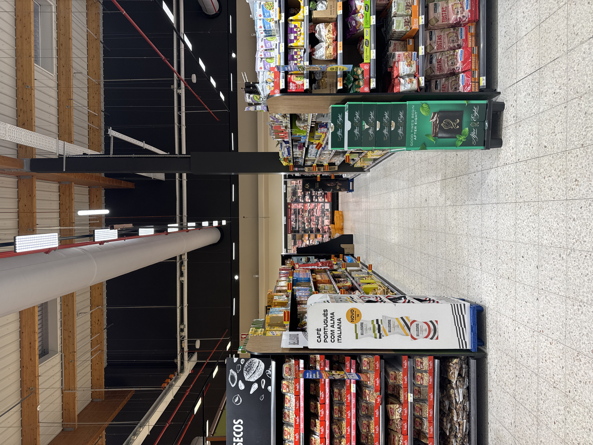 This is a low-angle, full shot of a grocery store aisle in Continente, located in the city of Castelo Branco, Portugal. The aisle is lined with shelves stocked with various food products. On the left, a display of nuts and dried fruits is prominent. To the right, shelves are filled with boxed cookies and snacks, and a display of "After Eight" chocolates stands out. The flooring is a speckled terrazzo. The overhead lighting is a mix of fluorescent and LED fixtures, with large white ventilation ducts running horizontally. The scene depicts a typical retail environment.