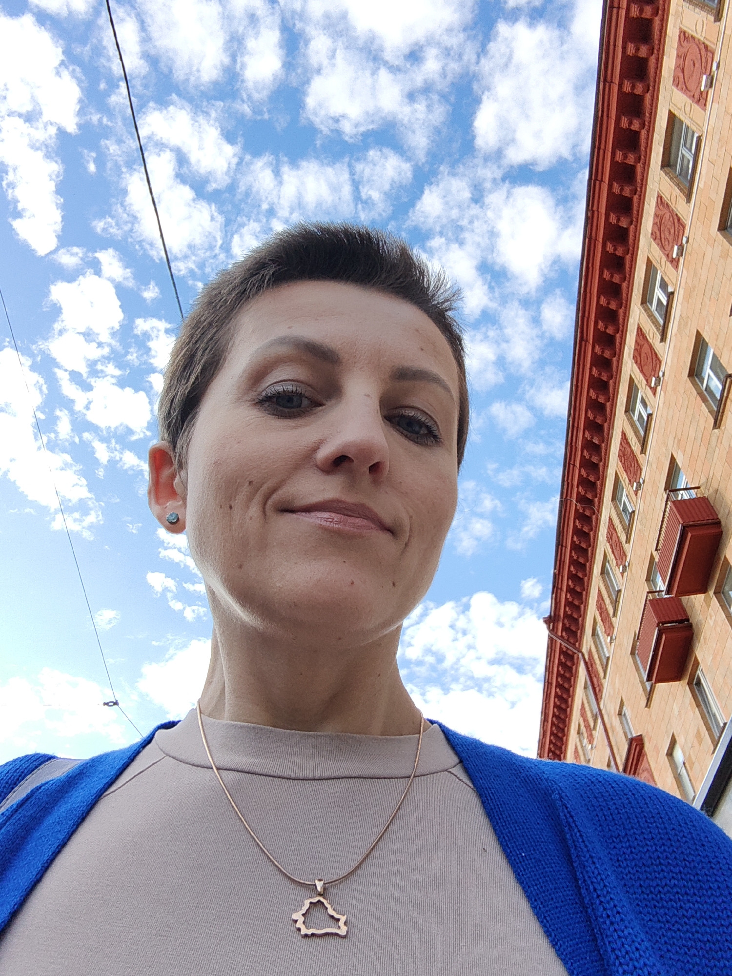 A low-angle, close-up, selfie-style photograph features a woman looking up at the camera. The woman has short, dark hair and is wearing a beige top and a bright blue knitted cardigan. She has a delicate gold chain necklace with a pendant shaped like the outline of Belarus. She is wearing a small, dark stud earring in her left ear. The background consists of a bright blue sky with fluffy white clouds, a dark power line, and a section of a building with orange brickwork and ornate red trim, characteristic of Stalinist Empire style architecture often found in Minsk, Belarus. The building has multiple windows and red balcony structures. The lighting suggests it is daytime.