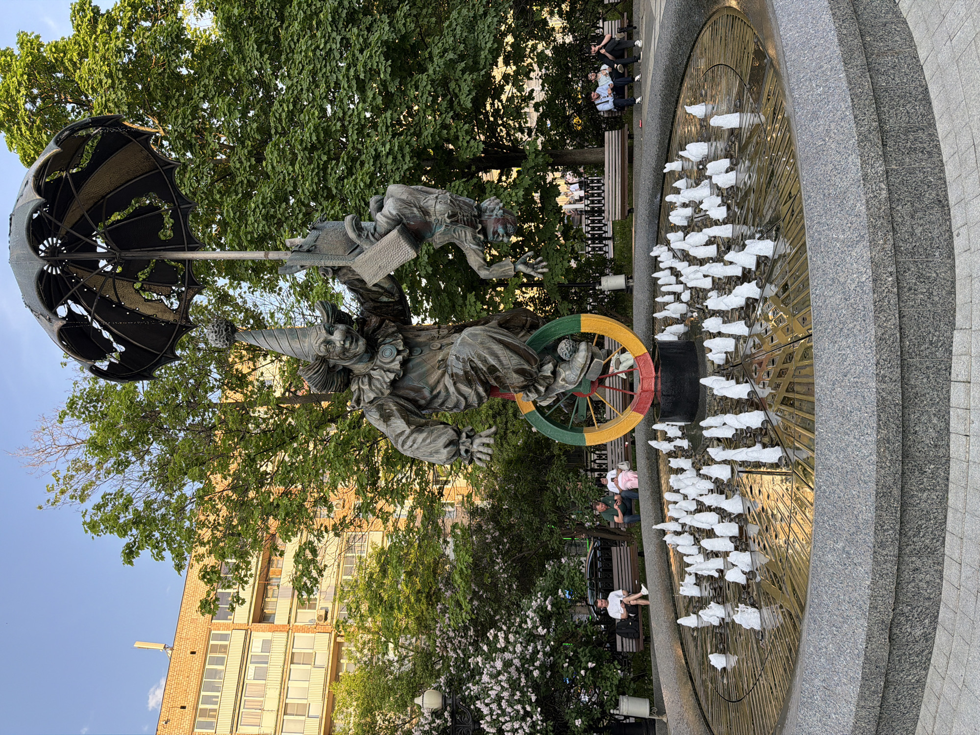 The image depicts the "Clown" sculpture in Moscow, Russia, situated in a public park. The sculpture features a clown with an umbrella overhead and another figure riding atop, next to a fountain with several water jets. People are sitting on benches in the background, enjoying the park setting. The backdrop includes trees and a multi-story building.
