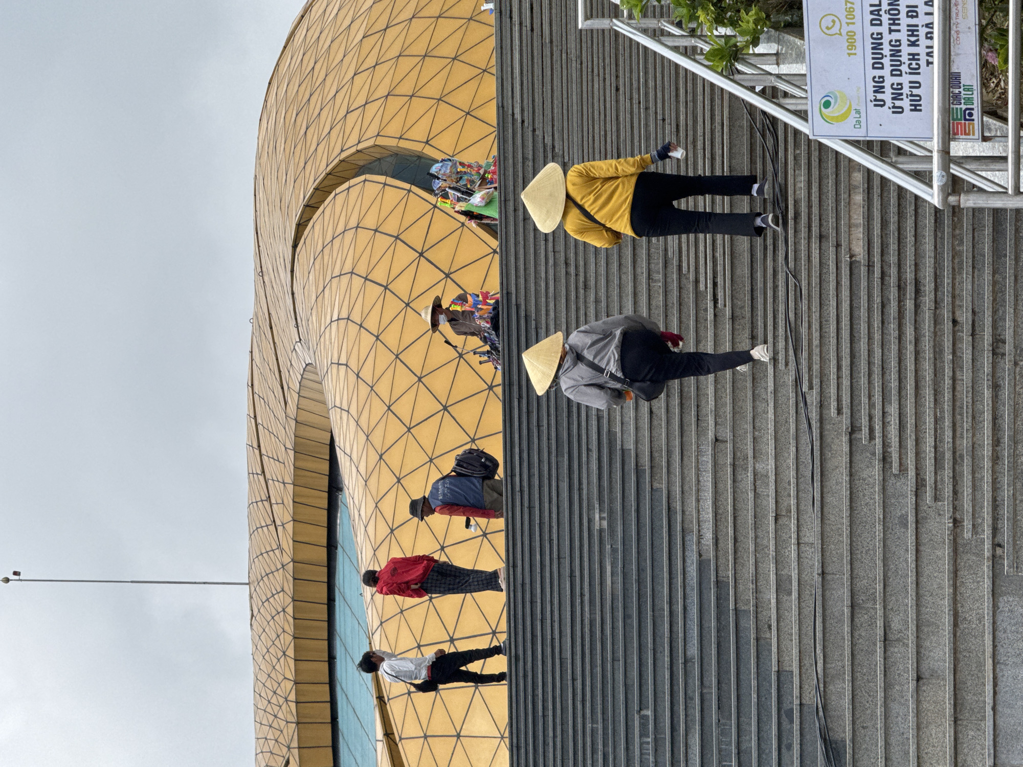 This image captures a wide outdoor scene in Da Lat, Vietnam, featuring a grand staircase leading up to a distinctive golden-domed building.
The central and upper portion of the image is dominated by a large architectural structure with an undulating, tessellated golden roof. The roof is composed of numerous geometric panels, possibly triangular, creating a textured, organic form with a prominent central crevice. A section of blue-tinted glass is visible beneath the golden roof. Above the building, a tall, slender pole with a small sensor or light fixture extends into a light grey, overcast sky.
The lower two-thirds of the image features a wide, multi-tiered staircase constructed of grey granite steps. A black cable runs horizontally across several steps in the mid-ground. On the far right, a silver metal handrail is visible, alongside a white and green sign with Vietnamese text, including "Da Lat" and "1900 1067," promoting a "Useful application when traveling to Da Lat."
Ten individuals are present in the scene.
On the lower section of the stairs, two adults wearing conical hats (nón lá) are ascending. The person on the left wears a grey top and dark pants, carrying a black shoulder bag. The person on the right wears a yellow long-sleeved top and dark pants, holding a small white object in their right hand.
Further up the stairs, nearer the top:
An adult male in a white long-sleeved shirt, black pants, and a black shoulder bag is walking upwards on the left.
An adult, seen from behind, wears a red long-sleeved top and dark patterned trousers, standing still.
An adult male in a blue and red vest over a dark shirt, dark pants, and a dark cap is standing with a black backpack, holding a white object in his right hand.
At the top of the stairs, two vendors are positioned at the base of the golden building. One vendor, on the left, wears a straw hat and face mask, surrounded by a display of small toys and pinwheels. A second vendor, on the right, is partially obscured behind a similar display of colorful merchandise.