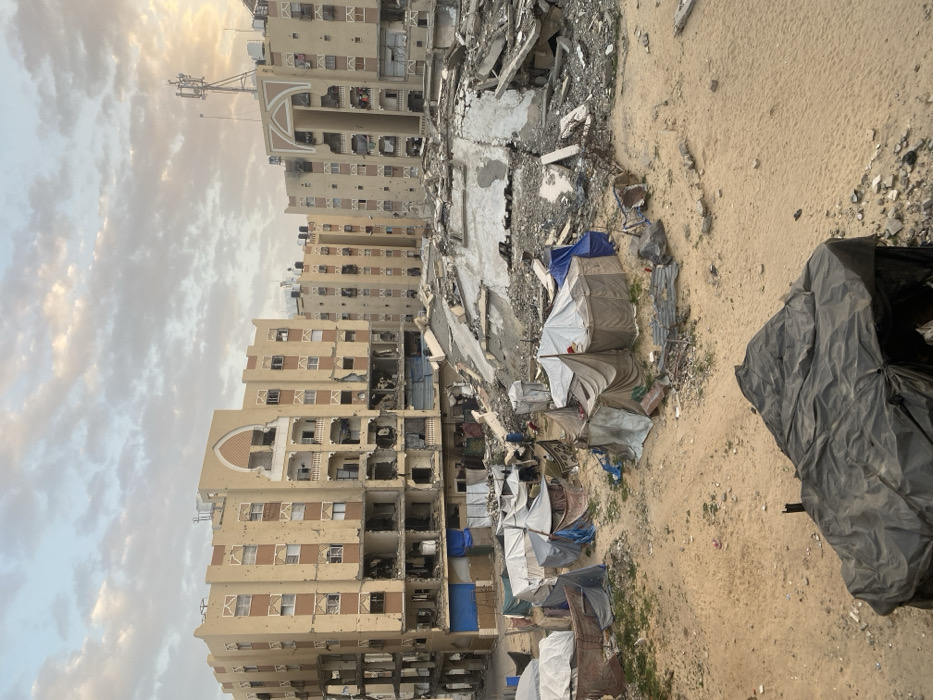 The image depicts a scene of significant destruction and makeshift living conditions under a cloudy sky. The photograph is oriented vertically, showing buildings and rubble on the left, and sandy ground with temporary shelters on the right.
In the background and extending into the mid-ground on the left, several multi-story buildings are visible. These buildings appear to be residential or commercial structures, constructed from light-colored, possibly beige or pale yellow, concrete or stucco. They show extensive damage, with numerous holes, missing wall sections, and charred areas, indicating past explosions or fires. Many windows are missing, and the interiors of some rooms are exposed, revealing debris and structural damage. The architectural style includes some decorative elements, such as arched shapes above what appear to be entranceways or balconies on the lower floors of some buildings. The highest building on the left has communication antennae or similar structures on its roof.
Between the damaged buildings and the foreground, there is a large expanse of rubble and debris, suggesting that other structures once stood there but have been reduced to ruins. Concrete slabs, twisted rebar, and assorted rubble are scattered across this area.
In the mid-ground and extending towards the foreground on the right, a cluster of makeshift shelters or tents is set up on sandy, uneven ground. These tents are constructed from various materials, including tarpaulins (some white, some blue, some gray), fabrics, and possibly corrugated metal sheets. They are small and appear to be temporary dwellings. Some have visible poles or frames supporting them.
The ground in the foreground and on the right side of the image is mostly barren, sandy earth with some sparse patches of dry grass or weeds. In the far right foreground, a large, dark-colored (possibly black) tarp or sheet is draped over something, partially obscuring it.
The sky above is filled with light-colored, stratocumulus-like clouds, with hints of warm, possibly golden or orange, light breaking through on the horizon, suggesting either early morning or late afternoon. The overall atmosphere conveyed by the image is one of devastation, displacement, and hardship. No discernible text is visible in the image.