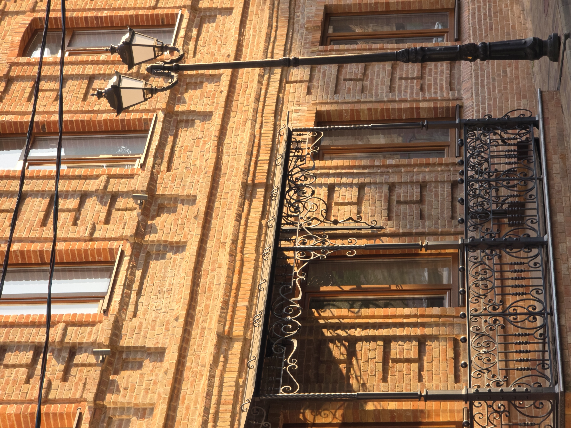 The image shows the facade of a brick building with ornate ironwork on a balcony and a lamppost in front of it. The building is made of red brick with decorative brickwork above the windows. The balcony features a metal canopy and a railing with intricate scrollwork. A double-headed lamppost with decorative lanterns stands to the right of the balcony. Shadows from overhead wires are cast on the building's facade. The scene is set during daylight with warm lighting, suggesting a sunny afternoon. The overall impression is of an older, possibly historic, urban architectural style. This building is located in Rostov-on-Don, Russia, specifically on Budennovsky Prospekt.