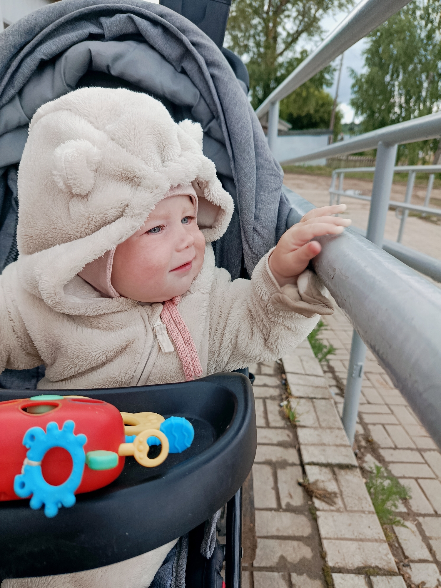 A baby in a stroller, wearing a beige fleece hooded jacket with ears, is reaching out and touching a silver metal railing. The stroller has a black tray with a red and blue toy. The location is an outdoor area in the city of Фоки, Russia, with a brick-paved ground, a white building, trees, and the railing.