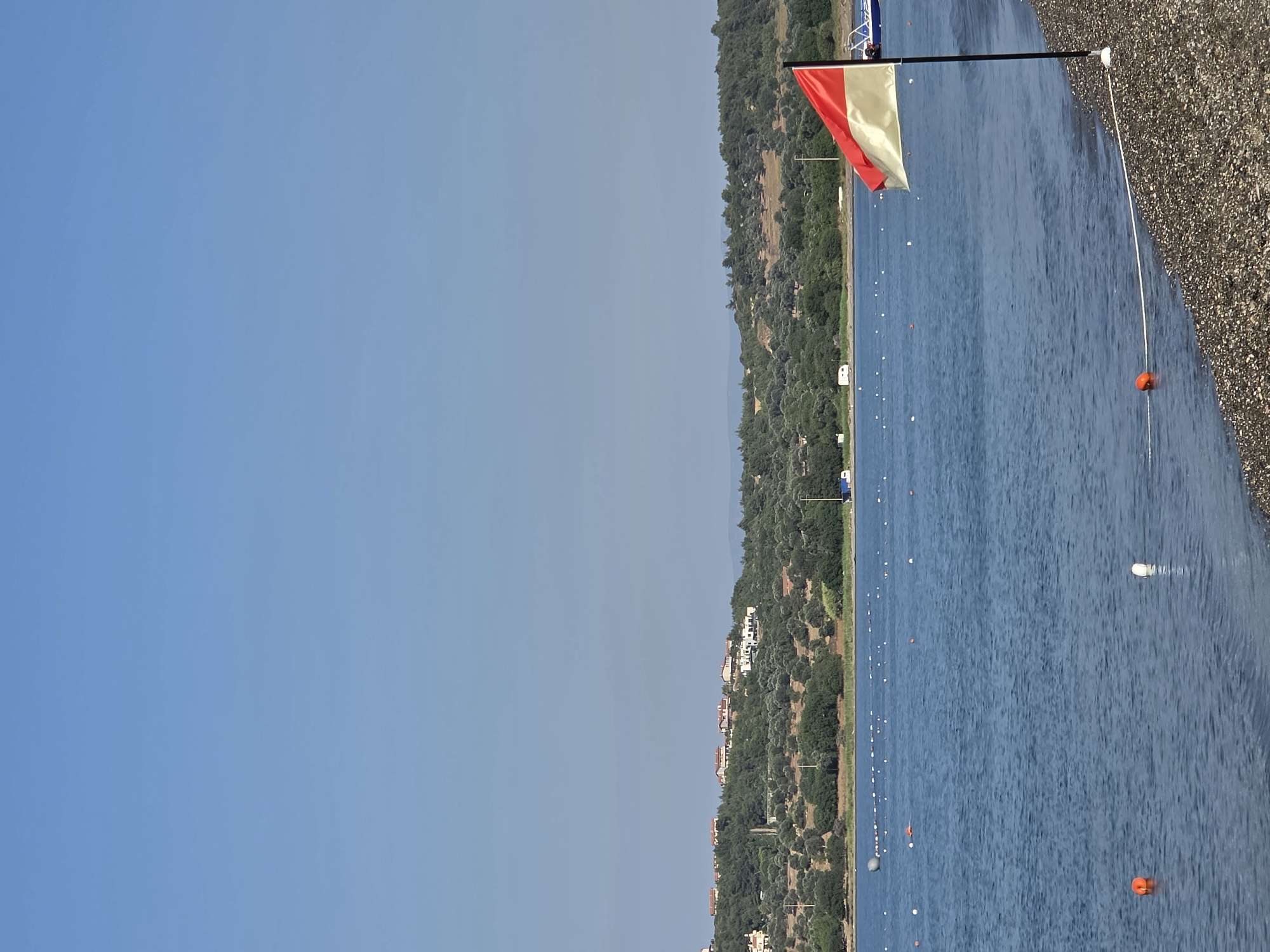 The image depicts a calm, blue sea with a gently sloping, pebbled shoreline in the foreground. A black pole stands on the shore, flying a red and white flag. Orange and white buoys are visible in the water, marking an area. In the background, a lush green hillside is dotted with houses and olive trees, suggesting a Mediterranean landscape. The sky is clear and blue. The scene appears to be a beach or coastal area in or near Seferihisar, Turkey, given the architectural style of the houses and the prevalence of olive groves. There are no people visible in the image.