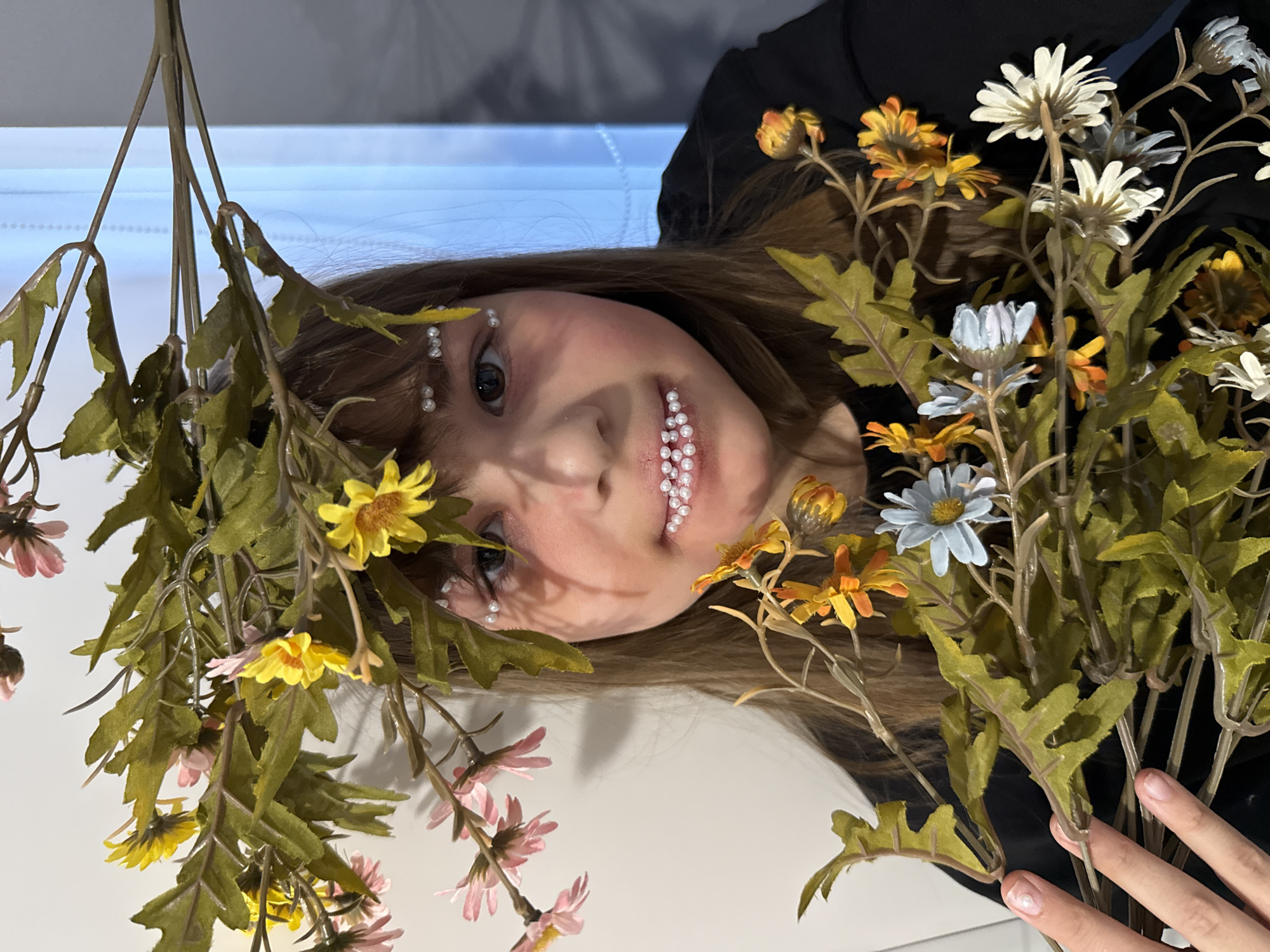 This media file presents a close-up portrait of a young woman, rotated 90 degrees clockwise, so her head is towards the left of the frame. She looks directly at the camera with a neutral expression.
The woman has light brown hair, which is partially covered by a dense arrangement of artificial flowers and foliage. Her face is adorned with small, white, pearl-like beads: a string of them trails above her left eyebrow, another cluster is placed at the outer corner of her left eye, and a significant number are applied over and beneath her lower lip. Her left eye is visible and dark, while her right eye is mostly obscured by the flowers.
She is surrounded by an abundance of artificial flowers of various colors, including bright yellow, delicate pink, crisp white, soft light blue, and warm orange/rust, all with green leaves and stems. These floral elements frame her head and extend across much of the image. In the bottom right corner, a hand, likely the subject's, is visible, gently holding a bundle of flower stems.
The background is a smooth, light-colored surface, possibly a wall or floor, with a soft blue light or reflection visible in the upper part of the image, suggesting an indoor setting. The overall presentation indicates an artistic or styled photoshoot. There is no visible text in the image.