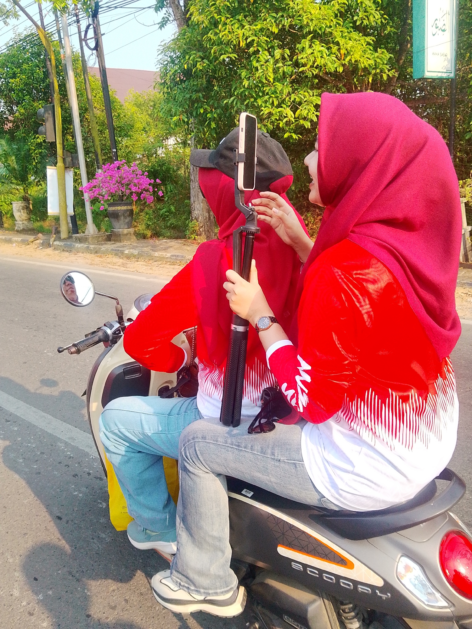 Two individuals, likely female based on attire and hairstyles, are seated on a scooter in what appears to be a semi-urban environment in Barabai, Indonesia. The person in the foreground, on the right, wears a maroon hijab and a red jacket with white vertical stripes on the sleeves. She is holding a selfie stick with a smartphone attached, facing towards the person behind her. The person in the back, partially obscured, also wears a maroon hijab and a red shirt, with a black baseball cap underneath. They are positioned behind the driver, with their hands near the selfie stick. The scooter, a white and grey model with "SCOOPY" branding visible, is parked on a paved road. The background features lush green trees, a building, and a street lamp with what might be traffic lights. A pot of vibrant pink flowers is visible on the left side of the image. The lighting suggests it is daytime, with sunlight casting shadows on the road.