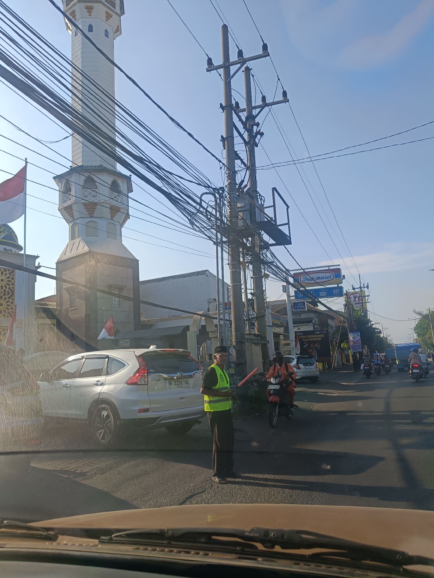 The image shows a street scene in Gedangan, Indonesia, on a clear, sunny day. A minaret of a mosque is prominently visible on the left, with intricate architectural details and a white and gold color scheme. A red and white Indonesian flag is flown near the mosque. A white SUV is parked on the left side of the road. A man in a bright yellow safety vest and a black cap stands in the middle of the road, holding a traffic signal baton, appearing to direct traffic. Several motorcycles are visible on the road, some carrying passengers. A blue truck is also present further down the street. Utility poles with a dense network of electrical wires stretch across the sky, obscuring parts of the buildings and the sky. Commercial signs, including one for "Indomaret," are also visible, indicating the presence of shops along the street. The road surface appears to be asphalt. The overall scene depicts a typical street environment with traffic and urban infrastructure in Indonesia.