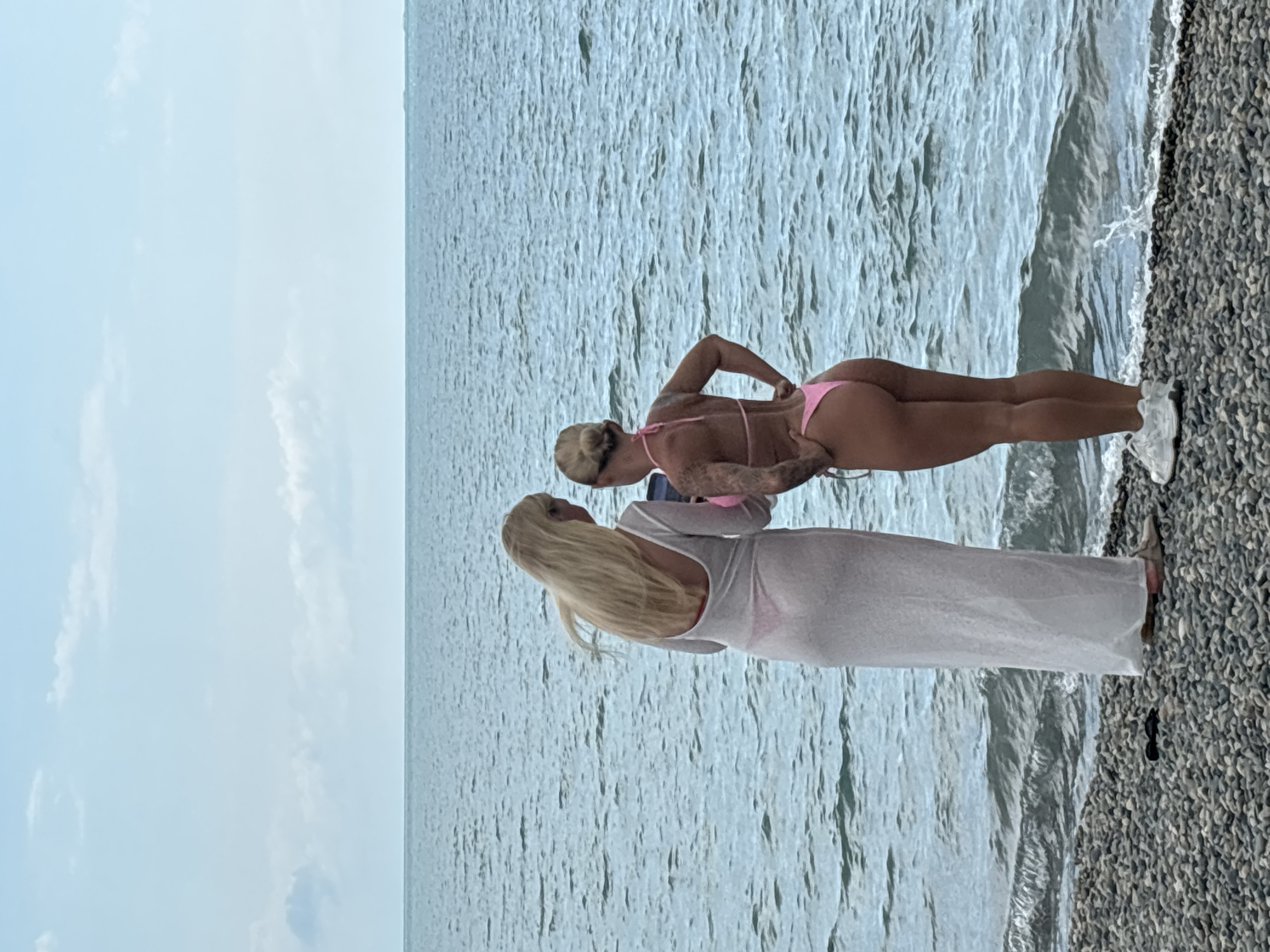Two women are standing on a pebble beach facing the sea. The woman on the left is wearing a long, sheer white dress over a pink bikini. She has long blonde hair and is looking down at a phone she is holding. The woman on the right is wearing a pink bikini and has her hair pulled back in a bun. She has tattoos on her arm and is looking at the phone as well. The sea is calm with small waves. The sky is overcast with a few clouds. This scene is likely in Sochi, Russia, as the question specified.