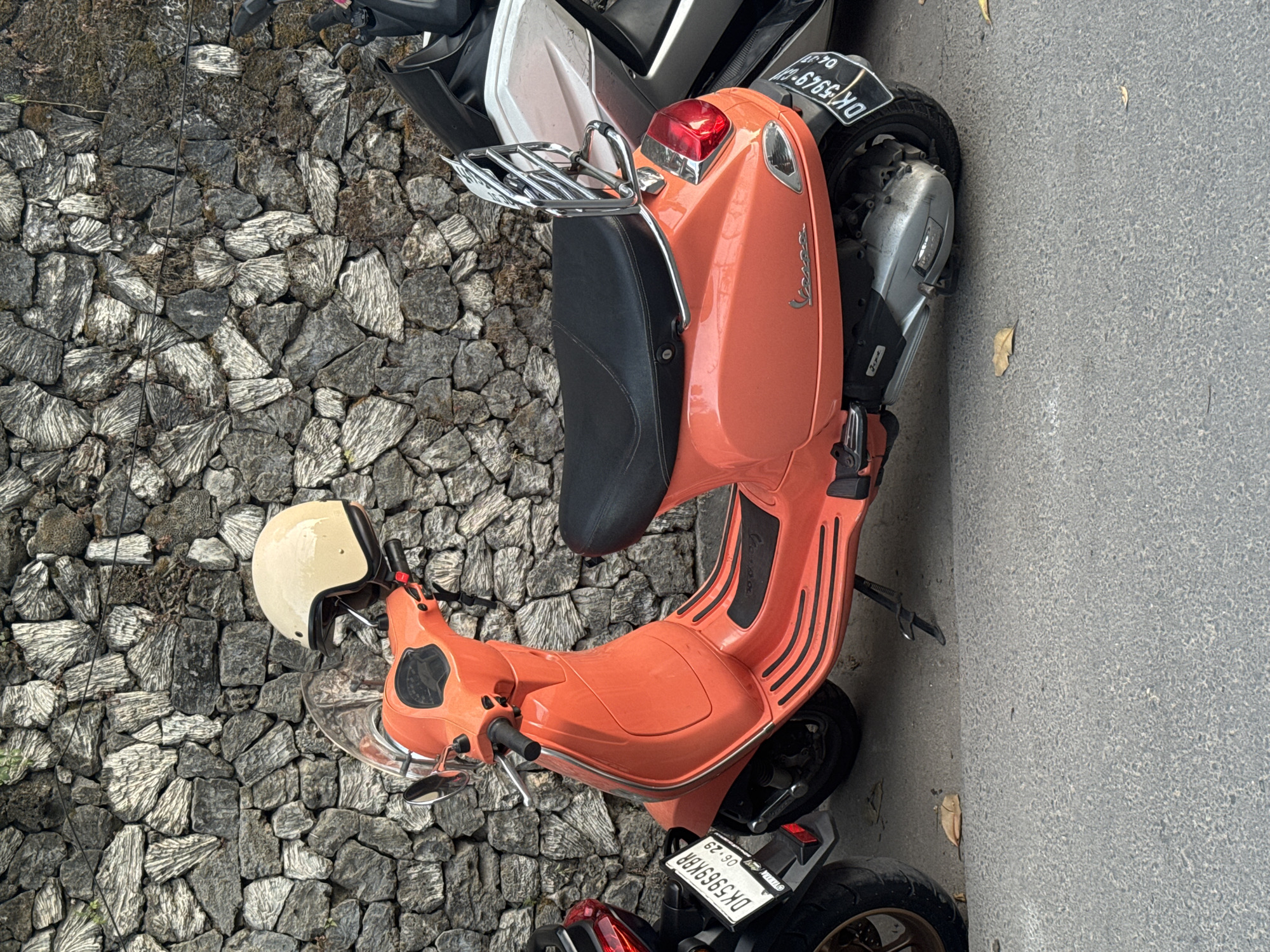 A coral-colored Vespa scooter, parked on a gray concrete surface, is positioned next to a rough, rock-filled area. The scooter is equipped with a black seat, a chrome rear rack, and a license plate that reads "DK 5949 XD." The Vespa is adjacent to another, partially visible motorcycle. A cream-colored helmet is placed on the front of the Vespa. Based on the license plate code "DK," the location is likely Denpasar, Indonesia.