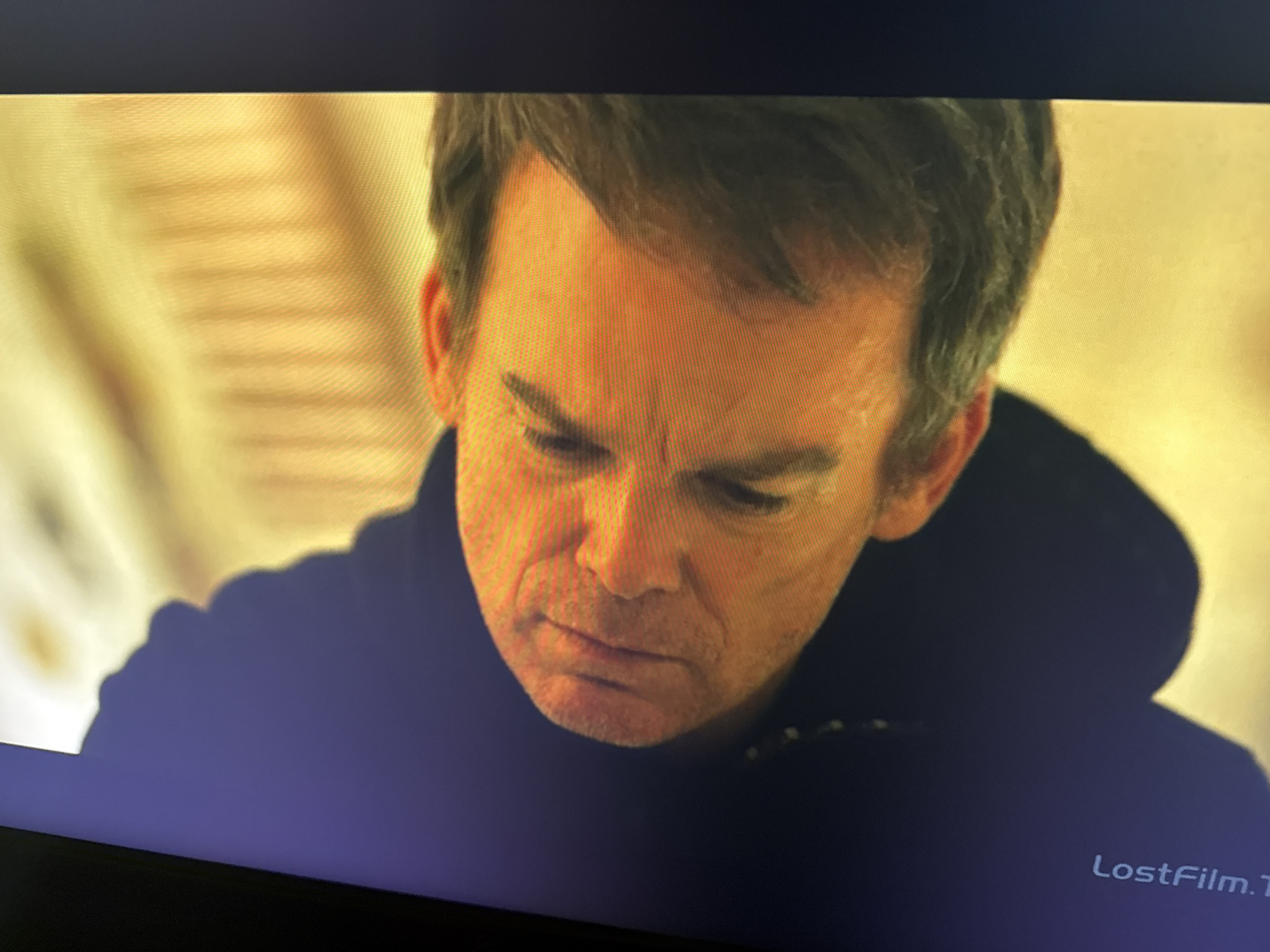 A medium shot depicts a fair-skinned man with short, greying hair, wearing a dark-colored hooded sweatshirt. His head is tilted downward, and he is looking down with a somber expression. The background is slightly blurred and features light-colored, possibly textured elements. The location is Unknown. A watermark "LostFilm.TV" is visible in the lower right corner of the image.