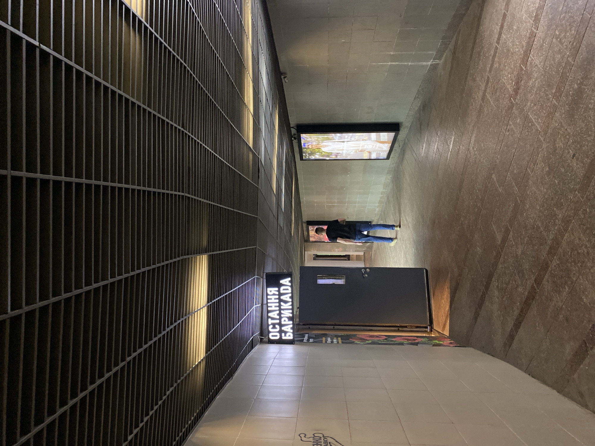 The media file shows an interior scene, likely a hallway or passage. The left side of the image is dominated by a dark, slatted metal structure, possibly a fence or a grated wall, with vertical light beams shining through from behind. The right side of the scene features a tiled floor and a stone wall.
In the background, a large rectangular screen is mounted on the ceiling, displaying a vibrant image. Further down the hallway, a person is visible standing near a closed dark door. The person is wearing a dark t-shirt and blue jeans, with their back to the camera.
A prominent illuminated sign with white text on a black background is positioned on the left side of the hallway. The text, in what appears to be Cyrillic script, reads "ОСТАННЯ БАРИКАДА". Below this sign, a colorful rug or mat is partially visible.
The lighting suggests an indoor environment, possibly at night or in a dimly lit area, with artificial lights creating highlights and shadows. The overall mood is neutral, with no strong emotional cues conveyed. The setting appears to be urban, perhaps a public space, a transit area, or an entertainment venue. The presence of the screen and the sign might indicate an advertisement or informational display.
