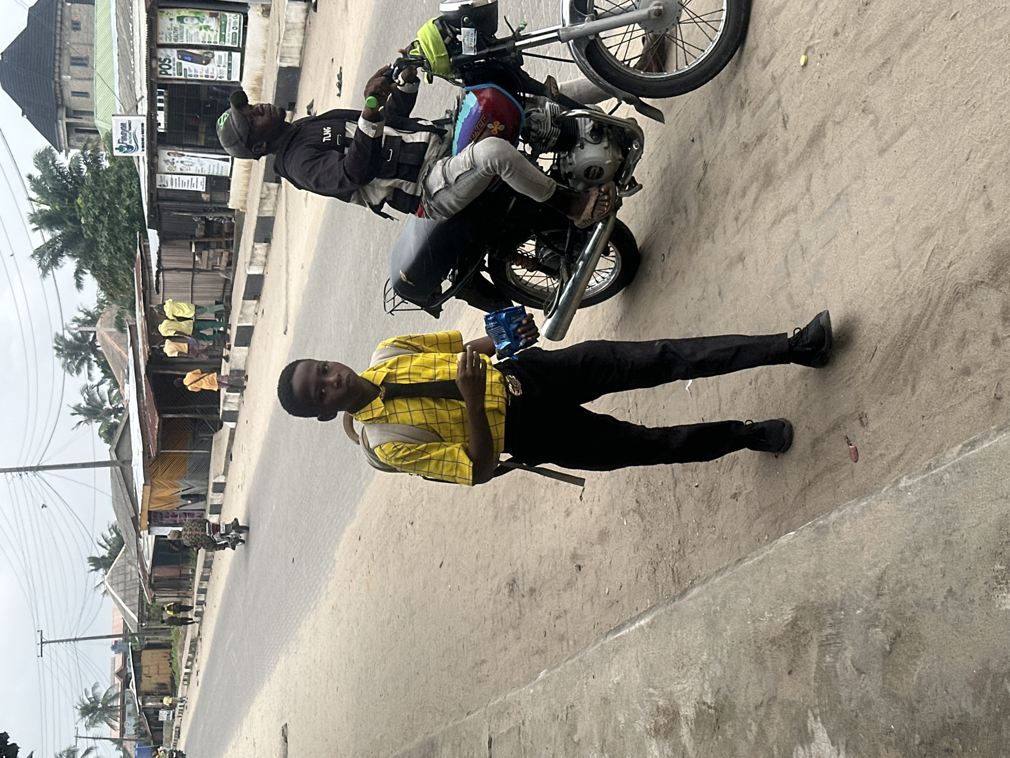 A medium shot captures a young boy in a yellow, checkered shirt, black tie, and black trousers with a backpack walking on a sandy road. He is holding a blue object in his right hand. To his right, a man on a black motorcycle is looking to his right. The motorcycle has a multi-colored design on its fuel tank. Behind them, several buildings with corrugated metal roofs line the street, some featuring yellow and brown siding. Palm trees are visible in the background, suggesting a tropical environment. Power lines and poles are also present. The scene is likely set in Badagry, Nigeria, a coastal city known for its historical significance and vibrant street life.