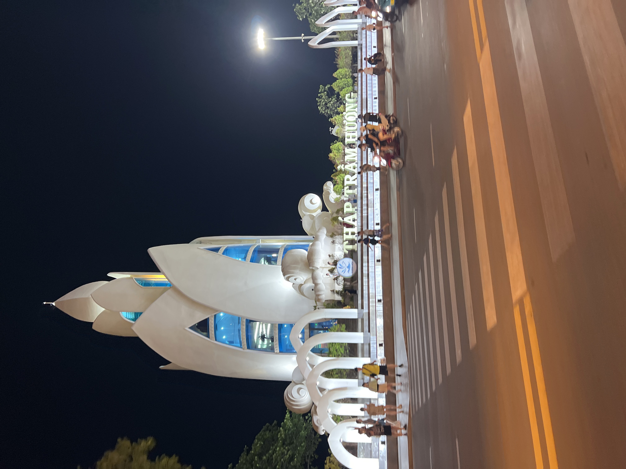 Here is a description of the image:
The image is a nighttime shot of the Lotus building in Nha Trang, Vietnam. The Lotus building, a white structure with blue windows designed in the shape of a lotus flower, is situated at the edge of a road. The road is marked with white pedestrian crosswalks and yellow road lines. Several people are walking along the road by the Lotus building. A street lamp is visible in the background. The sky is dark. The image is rotated.