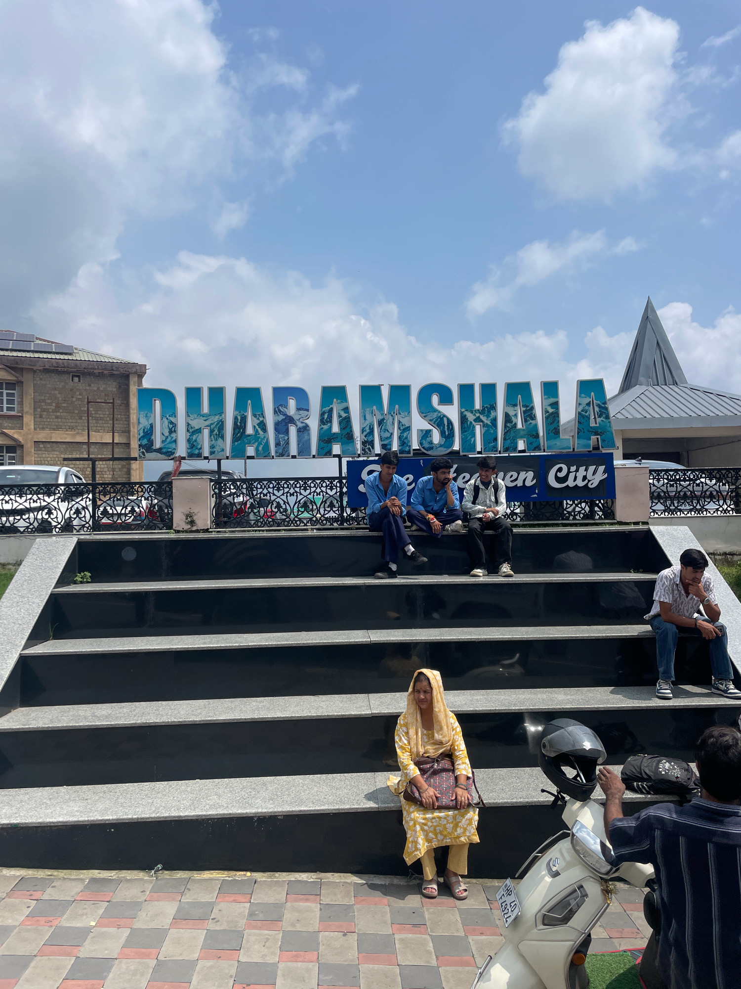The image shows the "DHARAMSHALA" sign in the city of Dharamshala, India. The sign is large and blue with a mountainous landscape filling the letters. Below the sign, a blue banner reads "The Heaven City". Several young men are sitting on wide, black, stepped platforms in front of the sign, some looking towards the camera and others looking away. A woman in a yellow outfit and headscarf is seated on the steps further down, with a bag on her shoulder. A scooter is parked in the foreground, with a helmet on its handlebars. A man in a dark striped shirt is standing next to the scooter, reaching towards the helmet. The sky is partly cloudy and blue. The scene appears to be a public gathering area or tourist spot.