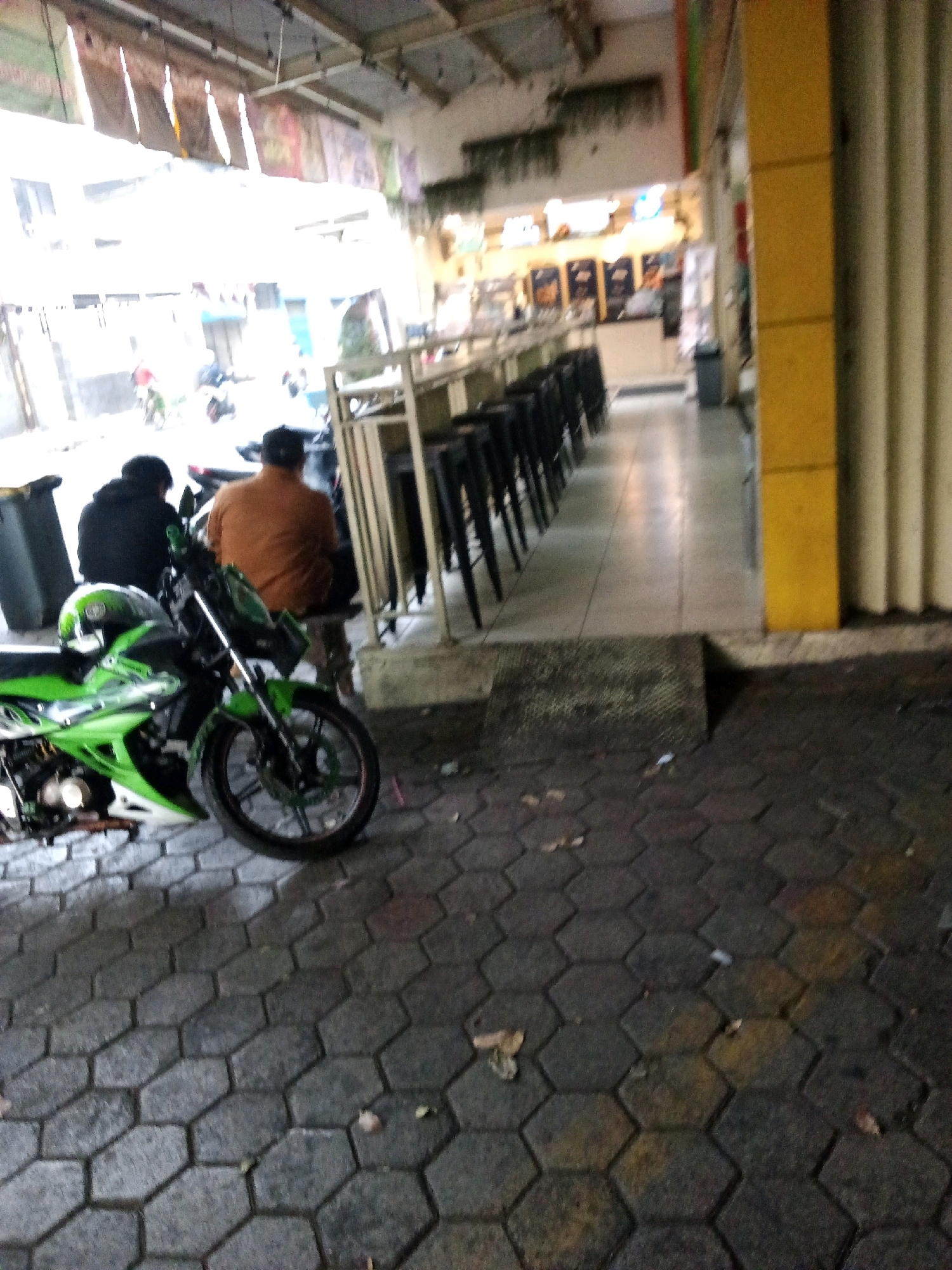 The image shows a street scene, likely outside a commercial establishment. In the foreground, a green and black motorcycle is parked on a patterned cobblestone pavement. Two people are seated on a bench behind the motorcycle, facing away from the camera. One person is wearing a black hoodie, and the other is wearing an orange shirt.
To the right of the motorcycle, a yellow pillar stands next to a corrugated metal wall. Further back, a row of tall black stools is arranged along a counter, suggesting a food or drink establishment. The background shows a walkway leading into a brightly lit interior with signage. The scene appears to be a public space in a city, possibly an area with street vendors or small shops. The overall lighting suggests it is daytime, but the light is diffused.