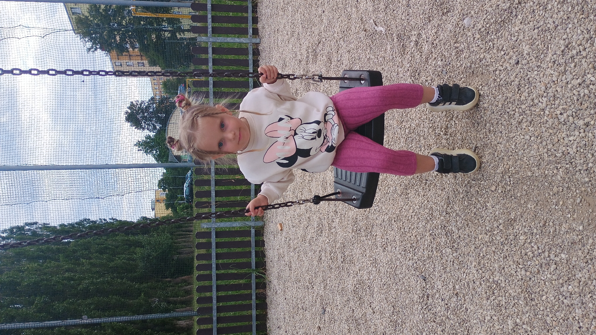 A young girl with blonde hair in two pigtails sits on a swing set in a playground. She is wearing a light-colored sweatshirt with a Minnie Mouse design and pink leggings. She has on black sneakers with white soles and velcro straps. The swing set is black with silver chains. The ground is covered with small light-colored gravel. In the background, there is a brown wooden fence, tall trees, and apartment buildings. The sky is overcast. The location is a playground in Sokolov, Czechia.
