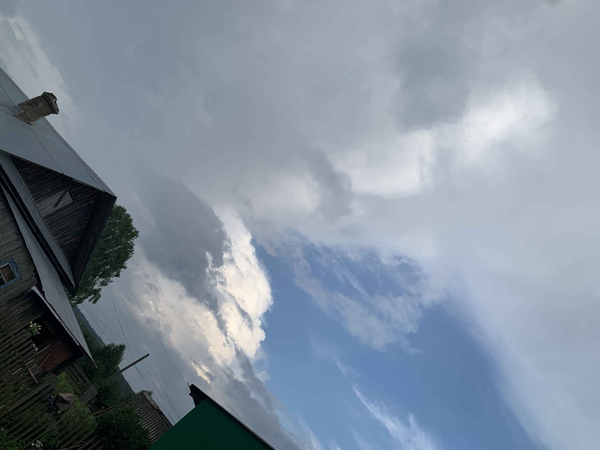 A low-angle, wide shot captures a dramatic sky over a rural scene in Panteleevka, Russia. The sky is divided, with a dense bank of dark, heavy clouds on the left, contrasting with a bright blue sky on the right, streaked with lighter clouds. In the foreground, the corrugated metal roof of a wooden house is visible, with a brick chimney. To the right, a portion of a bright green structure protrudes into the frame, also with a metal roof. Further back, another wooden house with a fence and trees are visible, with a hill in the distance. The lighting suggests it may be late afternoon or early evening, with the sunlight breaking through the clouds. There are no people or animals visible in the frame. The overall impression is one of a dynamic weather system passing over a tranquil, rustic landscape.