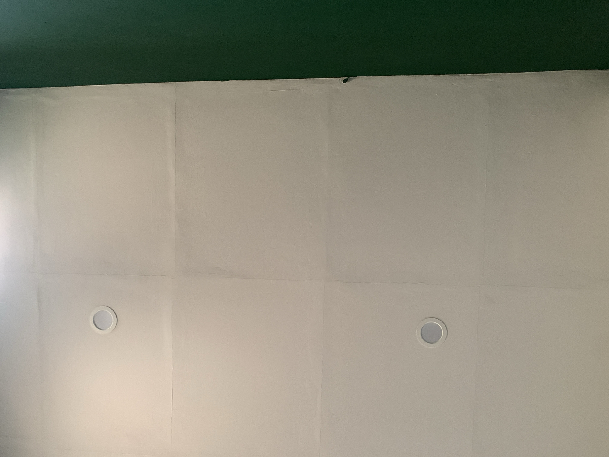 The image depicts an interior wall with two circular, recessed lights, one near the top and another near the bottom. The wall is painted a light, off-white color and appears to be divided into sections by faint horizontal and vertical lines, suggesting it might be a paneled or tiled surface. To the right of the wall, a portion of an adjacent surface painted a deep, matte green is visible. No people or specific objects that would indicate a particular location in the city of Kualadua, Indonesia, are present. The scene is static and appears to be a section of a building's interior.