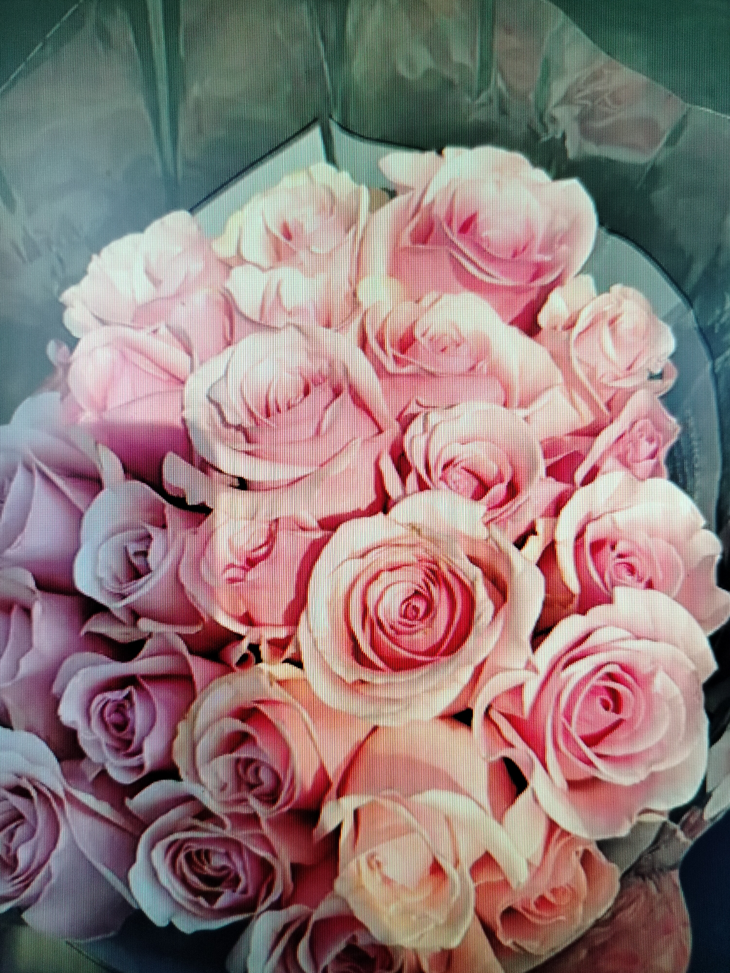 A close-up, overhead shot showcases a dense bouquet of pink roses, some fully bloomed and others in bud. The roses are tightly packed, their petals forming intricate swirls and layers. The bouquet is partially enclosed by dark green wrapping paper, which folds and drapes around the blooms. The lighting casts subtle shadows among the flowers, highlighting their textures and soft hues. No people are visible in the image. The image quality is somewhat degraded, possibly due to a low-resolution capture or compression artifacts, resulting in visible pixelation and a slight shimmering effect. No specific location in Yashkino, Russia, can be determined from the provided visual information.