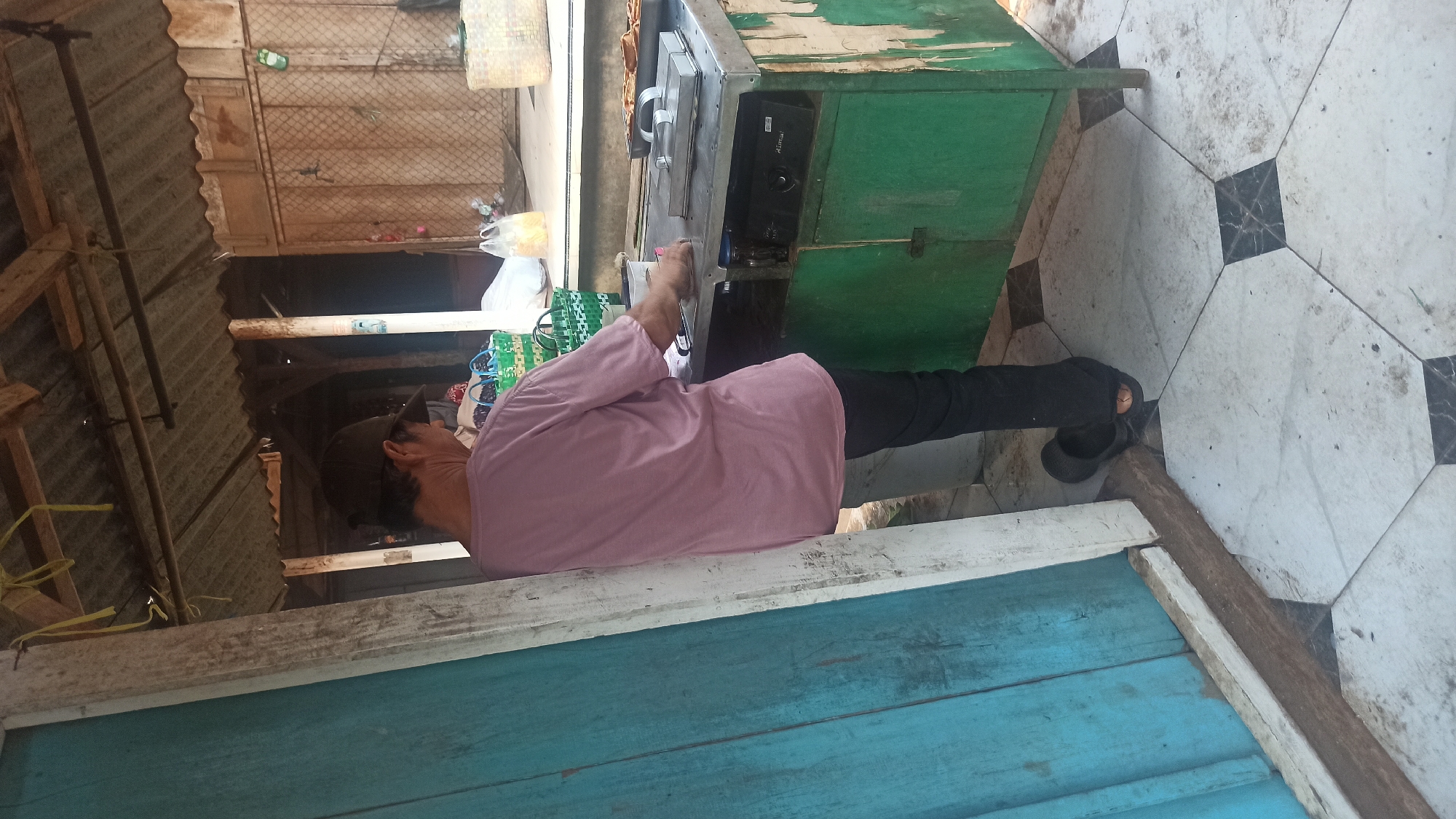 A person, viewed from the back, is standing at a food stall in Trisinar, Indonesia. The individual is wearing a pink t-shirt, black pants, and dark sandals. They are leaning against a blue wooden structure and appear to be preparing food on a metal counter. The stall has a green base and a metal top. On the counter, there is a gas stove, containers, and cooking utensils. In the background, there are woven baskets, plastic bags, and what appears to be a structure with a corrugated metal roof supported by wooden beams. The floor is tiled with a black and white geometric pattern.