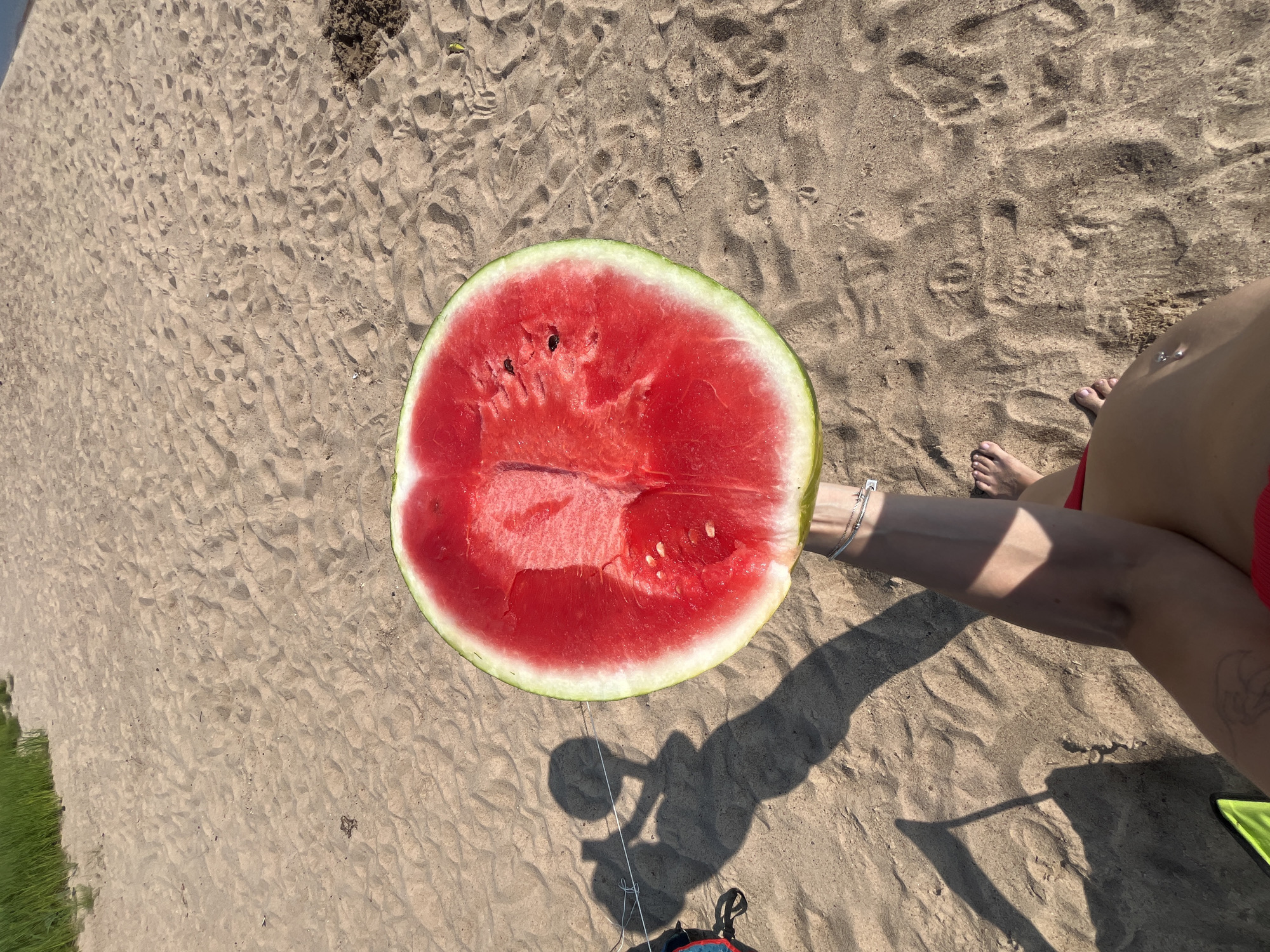 A slice of watermelon is held over a sandy beach, its bright red flesh and green rind contrasting with the light brown sand. The watermelon is positioned centrally in the frame, with its curved edge facing downwards. In the background, a shadow of a person holding the watermelon is cast on the sand, suggesting an outdoor setting. A thin string connects to something out of frame to the left, and a small patch of green grass is visible in the upper left corner. The sand has a textured appearance with visible footprints and small debris.