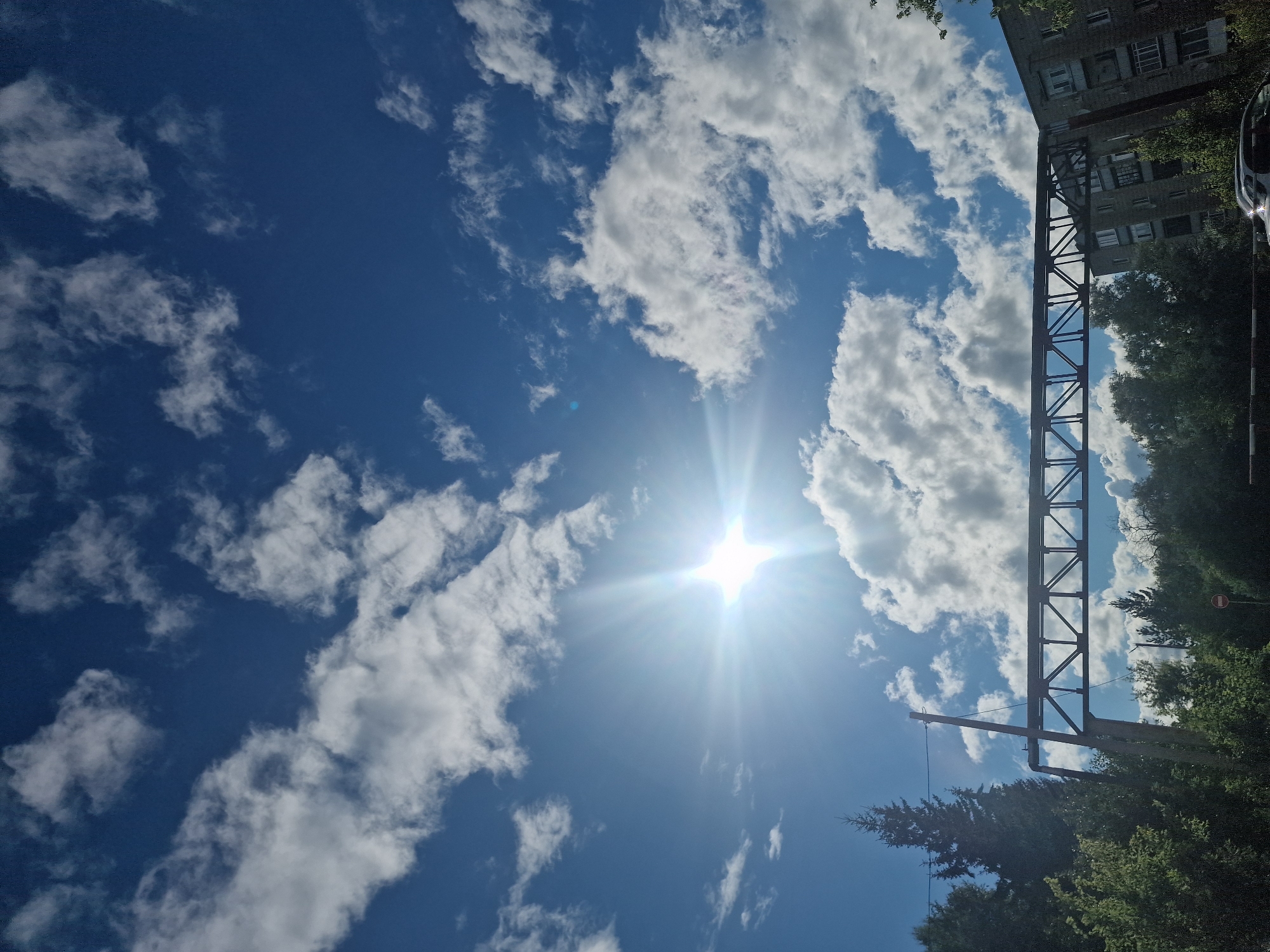 This media file captures a bright, sunny day with a clear blue sky dotted with fluffy white clouds. The sun is prominently visible, emitting strong rays that create a lens flare effect.
The scene includes elements of an urban or semi-urban environment. On the right side of the frame, a multi-story building with visible windows is present, suggesting a residential or office complex. Adjacent to the building and extending into the sky is a large, dark metal structure, possibly a part of an overpass, industrial equipment, or an elevated railway. Below this structure, dense green trees and foliage are visible, indicating a relatively natural or landscaped area within the built environment.
Further to the right, a dark-colored vehicle is partially visible, along with a black and white striped pole, which could be a road marker or part of a fence. A small red and white circular sign with a red horizontal bar is also visible on a pole amidst the trees, resembling a "no entry" or prohibition sign.
The image is oriented vertically, with the sky occupying the majority of the frame. The time of day appears to be midday, given the intense sunlight and the angle of the sun. The weather is clearly fair and sunny. There is no discernible text in the image. The overall impression is one of a typical sunny day in a developed area with a mix of modern architecture, infrastructure, and greenery. The location context of Kashgar City, China, suggests this scene is situated in that region.
