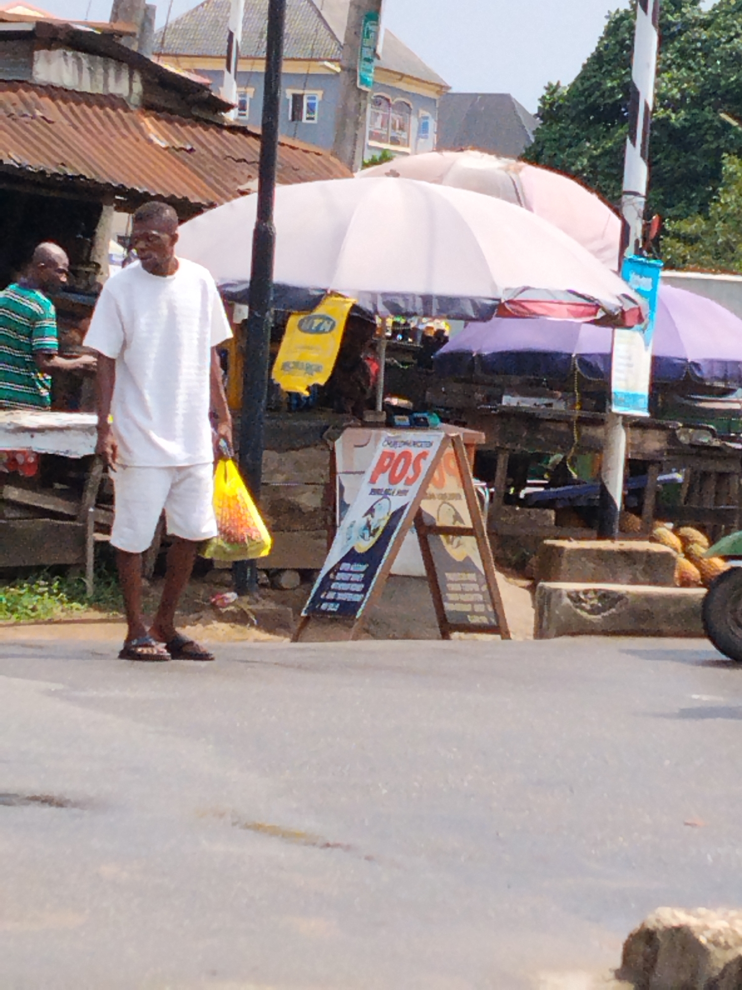 The image depicts a street scene in Aba, Nigeria. A man in a white shirt and shorts walks across the road carrying a yellow plastic bag. To his left, another man wearing a green striped shirt stands in front of a market stall with a corrugated iron roof. Further back, there are more market stalls covered by large white and purple umbrellas. A POS (Point of Sale) sign is prominently displayed. There's an MTN advertisement displayed on a yellow background. The street surface is asphalt, and the background includes buildings and trees. A pole with black and white stripes is visible. A wheel is partially visible at the right side of the image.