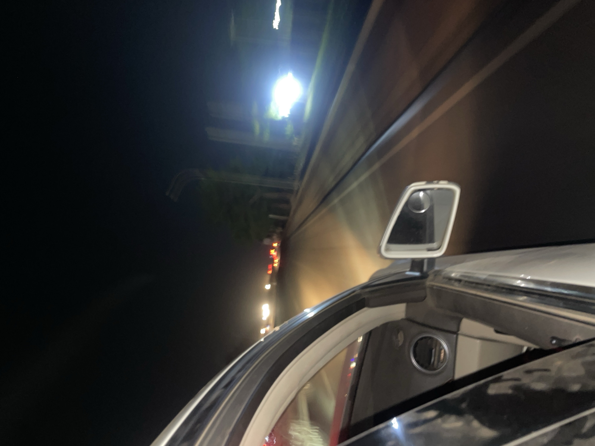 The image captures a nighttime scene from the perspective of a moving vehicle, looking out towards the right side. The top left half of the frame is dominated by darkness, with some faint, blurred reflections and distant lights. A prominent, bright light source, likely a streetlight or overhead illumination, is visible towards the top-center, casting a strong glow and reflections on the car's surfaces.
On the right side, a significant portion of a white or light-colored car is visible. This includes the vehicle's side-view mirror, which is in focus, and parts of the car's frame, windshield, and possibly the dashboard or interior. The car's metallic and glass surfaces reflect various light sources.
Further to the left and receding into the distance, a line of blurred red and white lights indicates other vehicles or structures along a road. The streaks and blurs in the lighting suggest the vehicle is in motion. The overall impression is that of driving on a road or highway at night. There are no people visible, nor any discernible text. The time of day is clearly night.