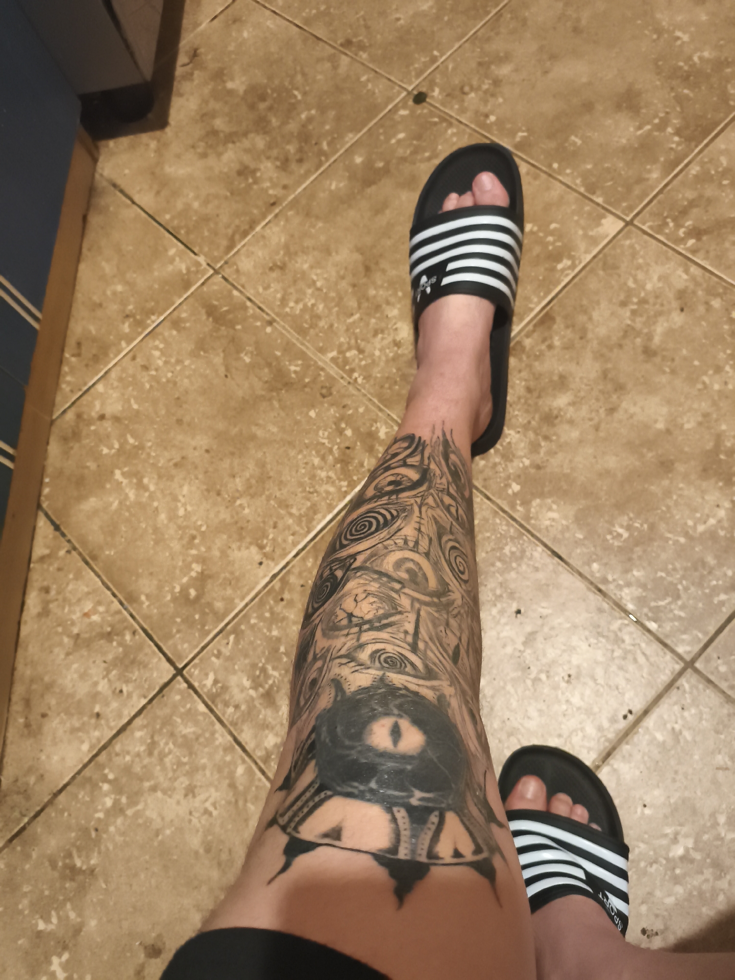 The image depicts a person's lower legs and feet against a backdrop of beige tile flooring. The person is wearing black and white striped Adidas slides. A prominent tattoo covers the majority of the person's left shin, featuring a large eye design and other intricate patterns. The location is potentially in Nowy Tomyśl, Poland, but no elements in the image confirms it. A portion of a dark cabinet with light-colored stripes is visible in the upper left corner of the frame.