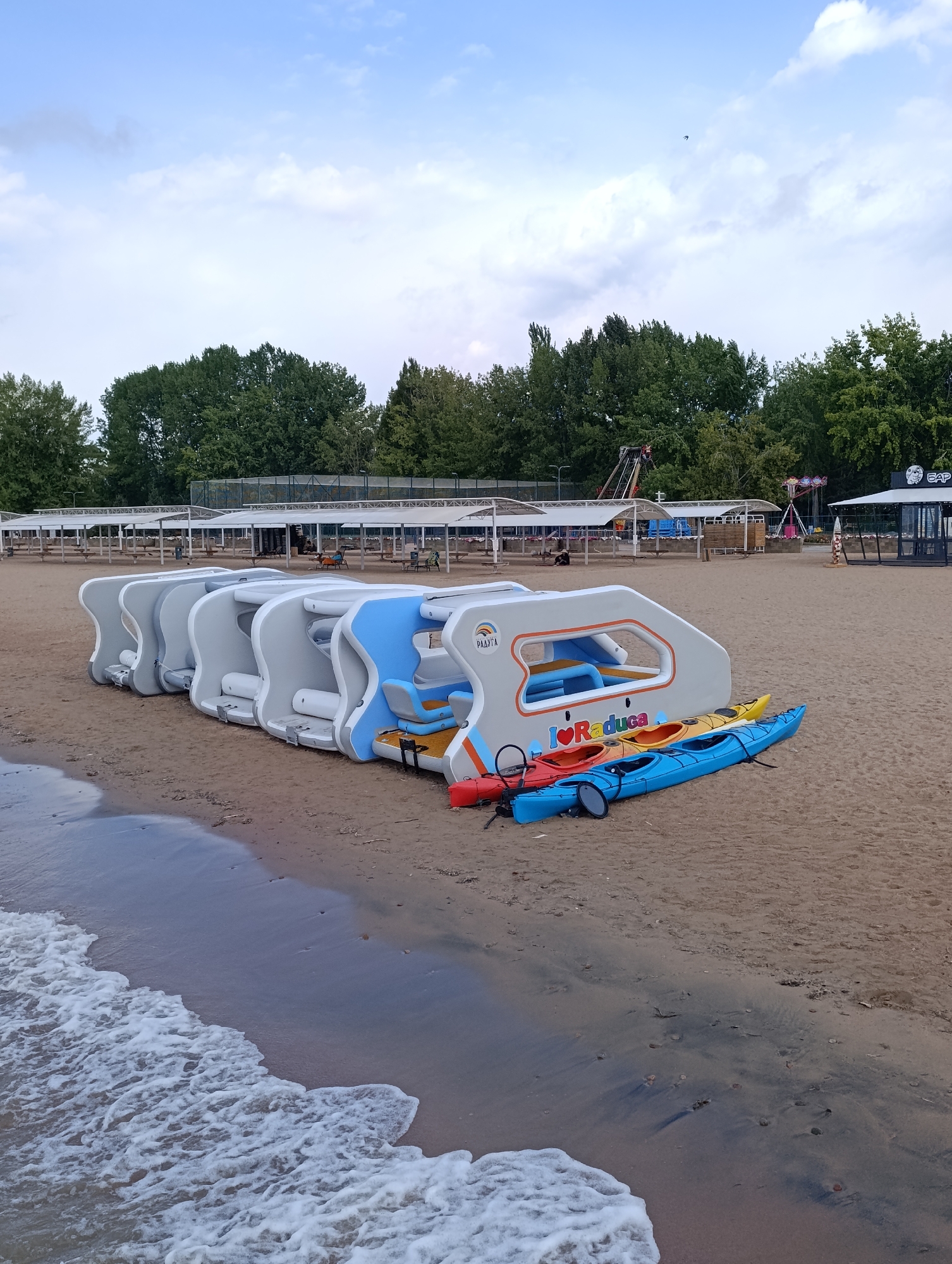 The image shows a row of empty, modular amusement park rides lined up on a sandy beach. The rides are mostly white with some blue accents. In front of the rides are three kayaks: one red, one orange, and one blue. The beach is located in Sary-Oy, Kyrgyzstan. The sky is cloudy and the ocean waves are visible in the foreground. In the background, there are trees and amusement park attractions.