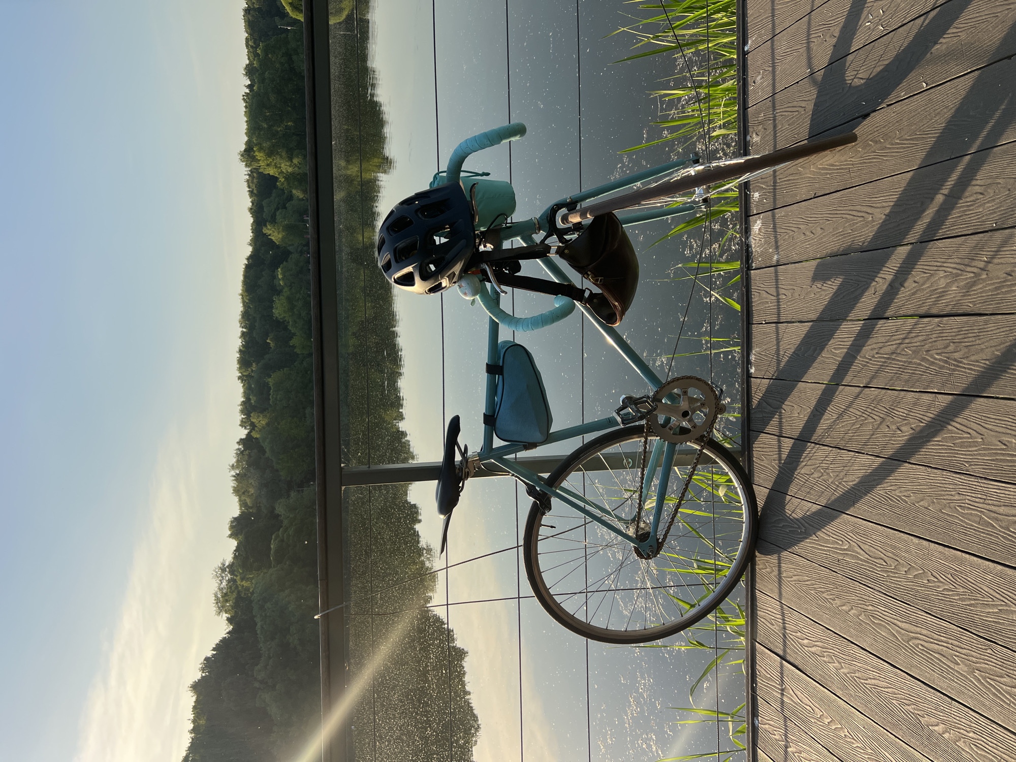 The image displays an outdoor scene featuring a light blue fixed-gear or single-speed bicycle parked on a wooden or composite deck surface. The bicycle faces to the right and leans against a dark-colored metal railing, which delineates the edge of the deck from a calm body of water.
The bicycle is equipped with drop handlebars wrapped in light blue tape, matching the frame. A black multi-vented cycling helmet rests on the handlebars, and a small, dark brown pouch or bag hangs from them. A small white bell is also attached to the handlebars. A light blue frame bag is secured to the top tube. The bicycle features a black saddle, a silver chainring, and thin black tires on silver rims.
The deck surface consists of planks with a simulated wood grain texture, oriented diagonally from the lower-left to the upper-right of the frame. Long, distinct shadows from the bicycle and railing are cast across the deck, extending towards the right, indicating that the primary light source, likely the sun, is positioned to the left. Small white flecks are scattered on the deck surface.
Beyond the railing, a placid body of water, possibly a lake or pond, reflects the bright, light blue sky and a dense line of green trees on its distant bank. The water's surface exhibits numerous small, bright specular reflections, particularly concentrated on the left side where direct sunlight appears to be hitting. Green, grass-like aquatic vegetation is visible along the water's edge, partially obscuring the lower section of the railing.
The background features a clear, light blue sky with a few faint, wispy white clouds, positioned above a continuous expanse of dense green deciduous trees, suggesting a forest or park environment. The overall lighting indicates either early morning or late afternoon. No individuals are visible in the image.
The precise location in the city of Unknown, Unknown, cannot be determined from the visual information provided.