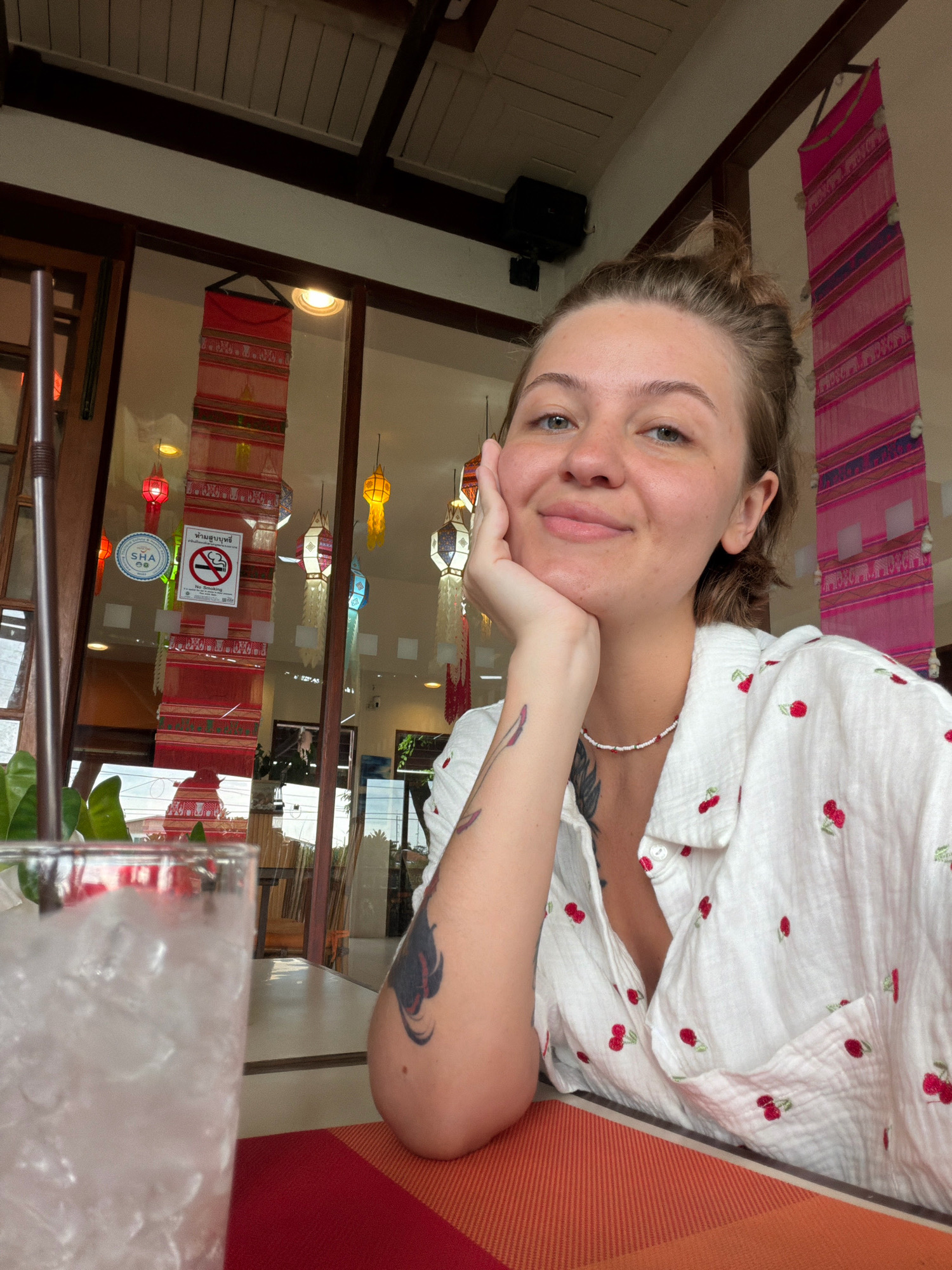 A woman is taking a selfie in a restaurant in Hua Hin City Municipality, Thailand. She is seated at a table with a glass of ice water in front of her. The woman has her hair tied up and is wearing a white shirt with a cherry pattern. She is resting her chin on her hand and smiling at the camera. In the background, there are windows with hanging lanterns and banners, indicative of a Thai restaurant. The scene appears to be during the daytime.