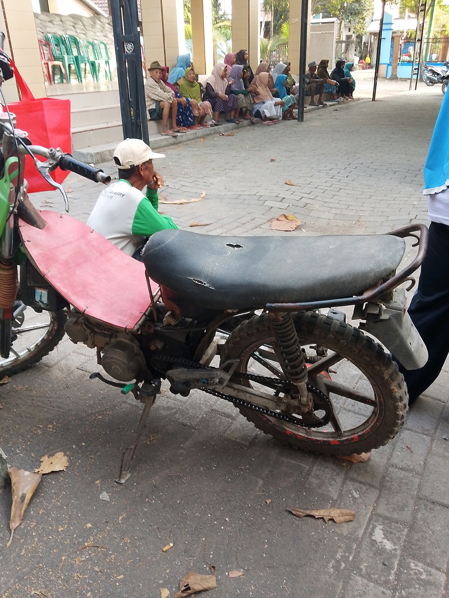 The image depicts an outdoor scene in Wadang, Indonesia, likely on a paved walkway or courtyard. In the foreground, a dirt-covered motorcycle is parked, its red seat cover and black seat clearly visible. The motorcycle's engine and rear wheel with a chain are also apparent.
To the left of the motorcycle, a person wearing a white shirt with a green collar and sleeves is seated, looking down and to the right, holding something in their hand. They are wearing a light-colored cap.
In the mid-ground, a group of individuals, predominantly women wearing headscarves, are seated on a raised step or bench. They are dressed in various traditional Indonesian attire. Behind them, several green and red chairs are stacked, suggesting a public gathering or event space.
The environment is characterized by a paved ground surface made of interlocking bricks, with scattered leaves and debris. The background reveals a building with columns and a tiled roof, consistent with Indonesian architecture. The overall scene suggests a casual, everyday setting in a public area.