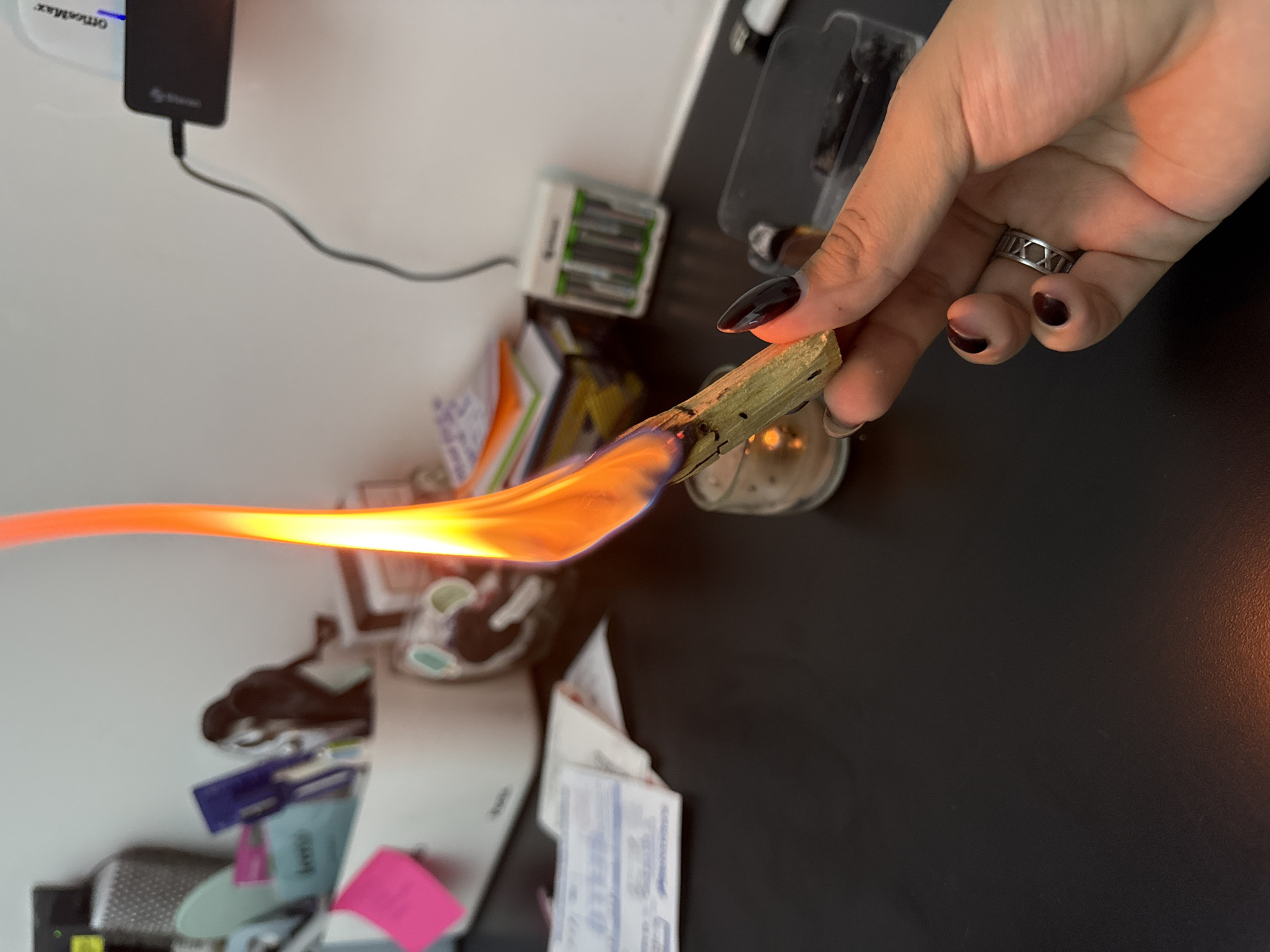 The image shows a person holding a burning piece of palo santo wood over a desk. The desk is dark and cluttered with various items, including a candle, batteries, notebooks, and other miscellaneous objects. The person's hand is visible, and they are wearing a ring. The flame is large and orange. The background is a white wall with some items attached to it. The location is not discernible.