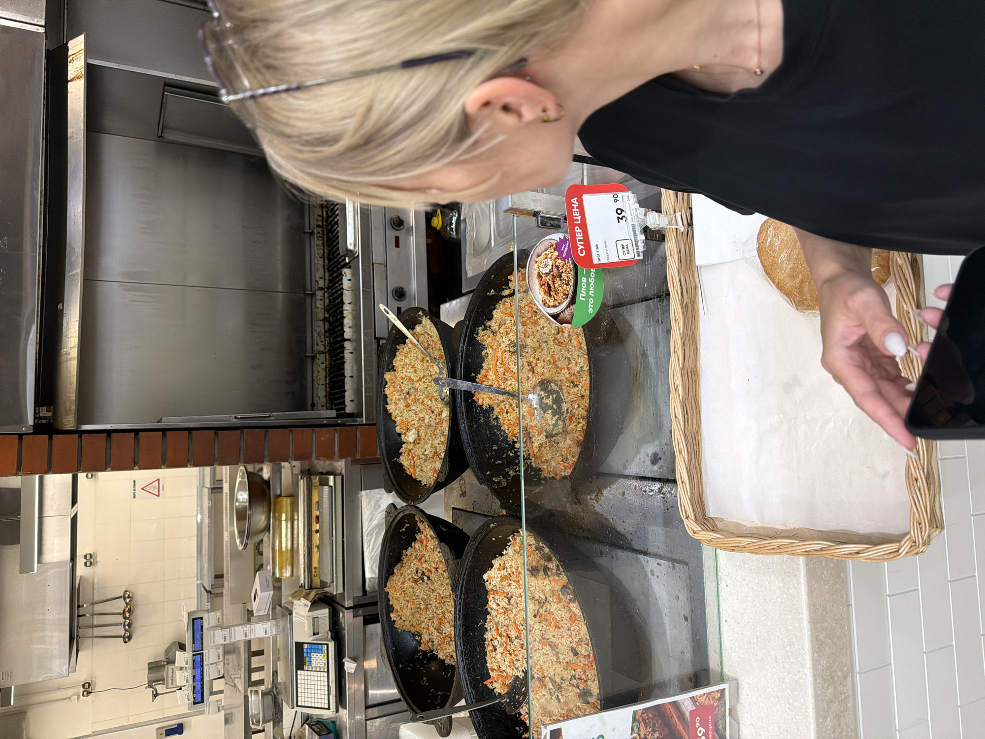 This media file captures a close-up of a woman with blonde hair, seen from behind and above, as she looks down at a food display. She is wearing a black top and appears to be in a self-service food area, likely a supermarket or cafeteria, with stainless steel kitchen equipment visible in the background.
The main focus is on several large, round, black cooking pots filled with pilaf. The pilaf appears to be a rice-based dish with visible ingredients like carrots and possibly meat. To the right of the pots, a woven basket is partially visible, containing a bread item wrapped in plastic.
A price tag is prominently displayed, indicating "СУПЕР ЦЕНА" (Super Price) and showing "39 90". Below this, another sign partially obscured reads "Плов - Это любоЕ" (Pilaf - It's everyone's favorite). The lighting suggests an indoor setting during daytime. The woman's posture and the surrounding context imply she is selecting food or making a purchase. The overall scene is casual and everyday, likely taking place in a grocery store or a food court within Nizhny Novgorod, Russia.