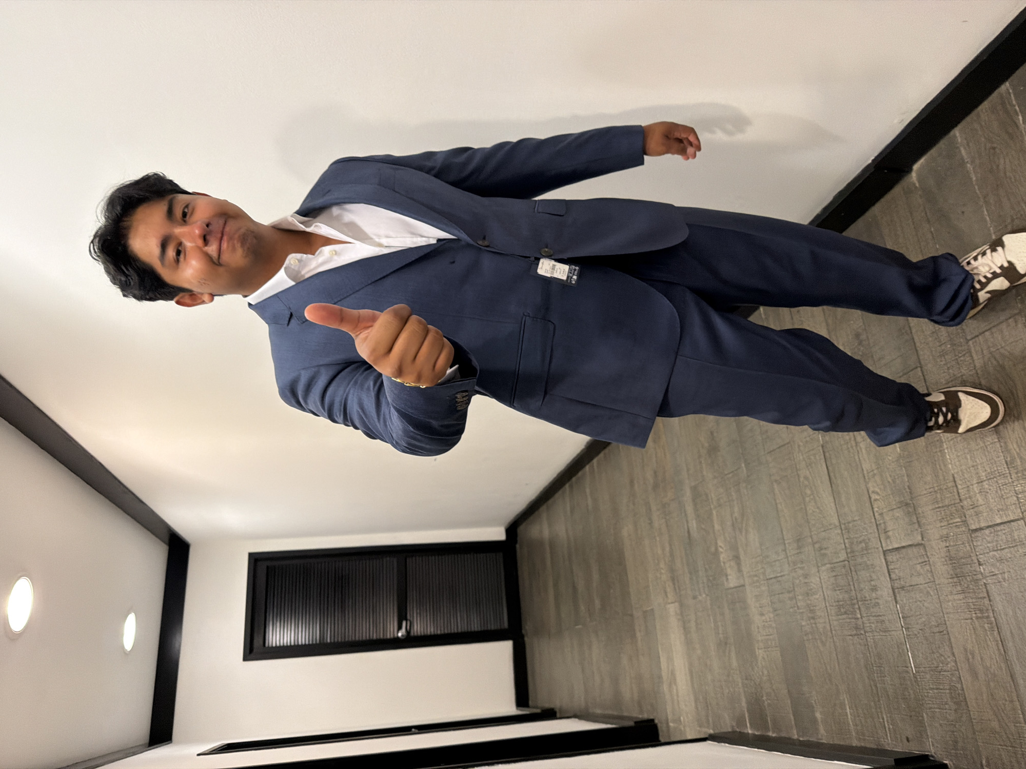 A young man stands in a hallway, dressed in a dark blue suit with a white dress shirt, and off-white sneakers with brown details. He is giving a thumbs-up with his right hand. The hallway has white walls with black trim. A window with vertical slats is visible on the left wall, along with two circular light fixtures. The floor is covered in gray wood-look planks. The location of the image is likely Puebla, Mexico.