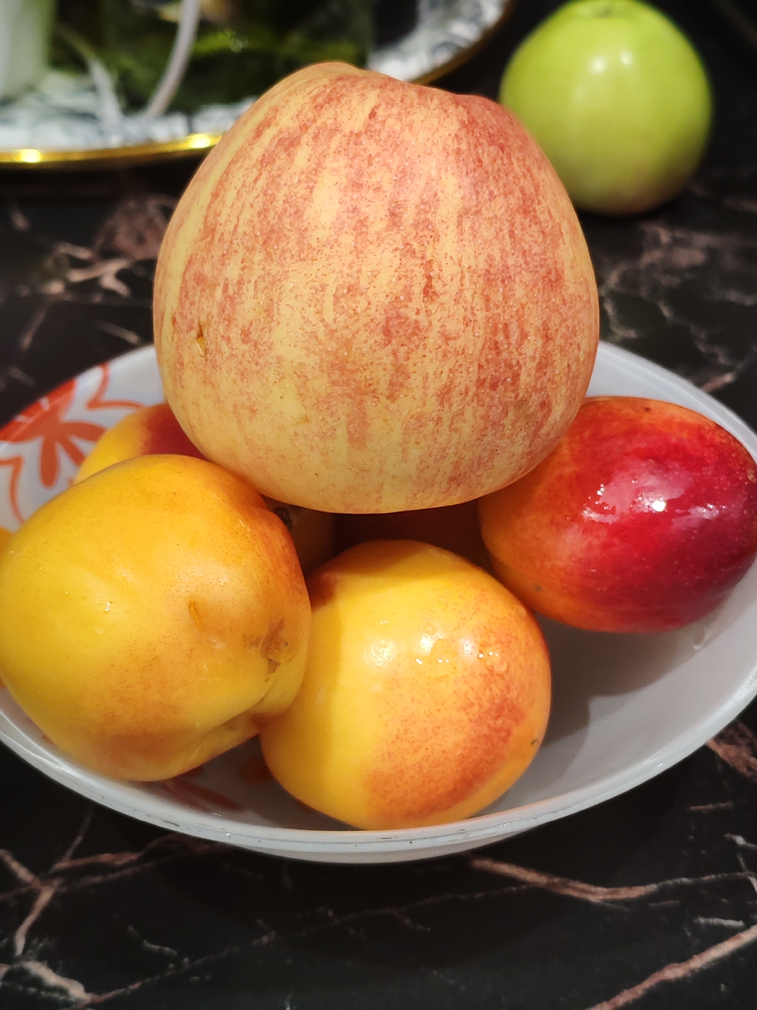 A still life image shows a white bowl filled with several fruits, including two yellow-orange nectarines in the foreground and a larger, red-striped apple on top, and a small red plum or nectarine to the right. Behind the bowl, a bright green apple sits on a dark, marble-like surface. The background is blurred, but appears to show more fruit and greenery. The lighting is dim, with some highlights on the fruit surfaces. The scene is likely indoors. There is no indication of a city or specific location.
