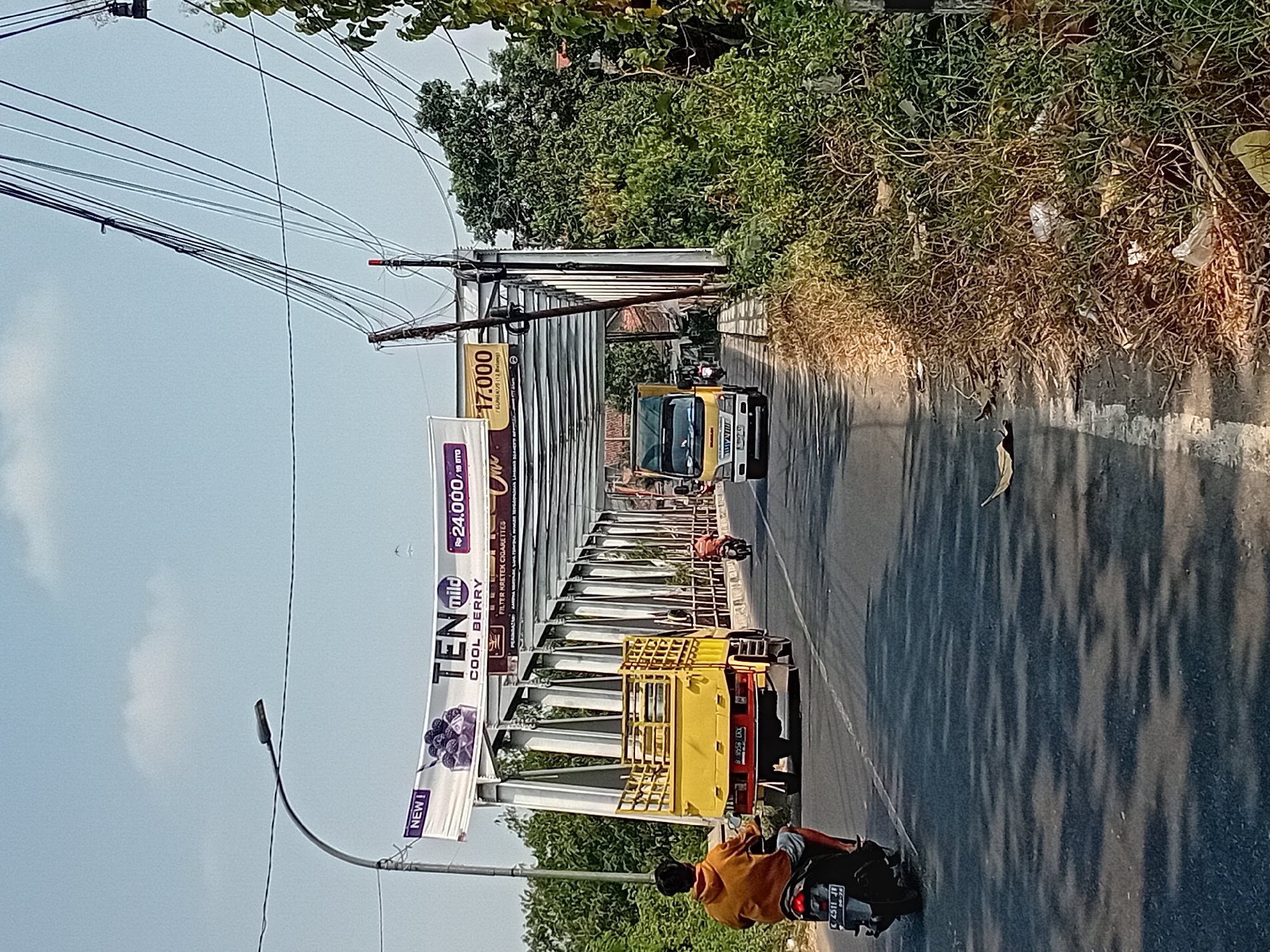 The image depicts a scene on a road in Gringging, Indonesia, likely a bridge, on a clear, sunny day. In the foreground, a person wearing an orange hoodie and shorts is riding a motorcycle. The motorcycle has a license plate with the characters "C 4511 JL". Ahead on the road, a yellow truck with a metal cage on its back is driving. Another motorcycle, ridden by a person in an orange shirt, is in front of the truck. A white truck with a logo on its side is further down the road. Overhead, a large banner advertising "TEN mild COOL BERRY" is suspended, displaying prices of "Rp 24.000" and "Rp 17.000". The bridge structure is visible, made of metal girders. The surrounding environment consists of lush green trees and vegetation, with some dry grass and scattered litter along the roadside. Power lines are prominently strung above the roadway and the bridge.