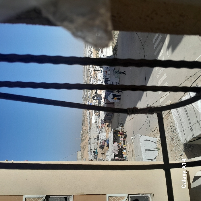 View from a window with metal bars looking out onto a densely packed urban or refugee camp setting. Makeshift structures and tents populate the landscape, with a road running through the middle marked by a zebra crossing. Figures are seen walking along the road. The sky is clear and blue. The lower portion of the image shows part of a building with visible windows.